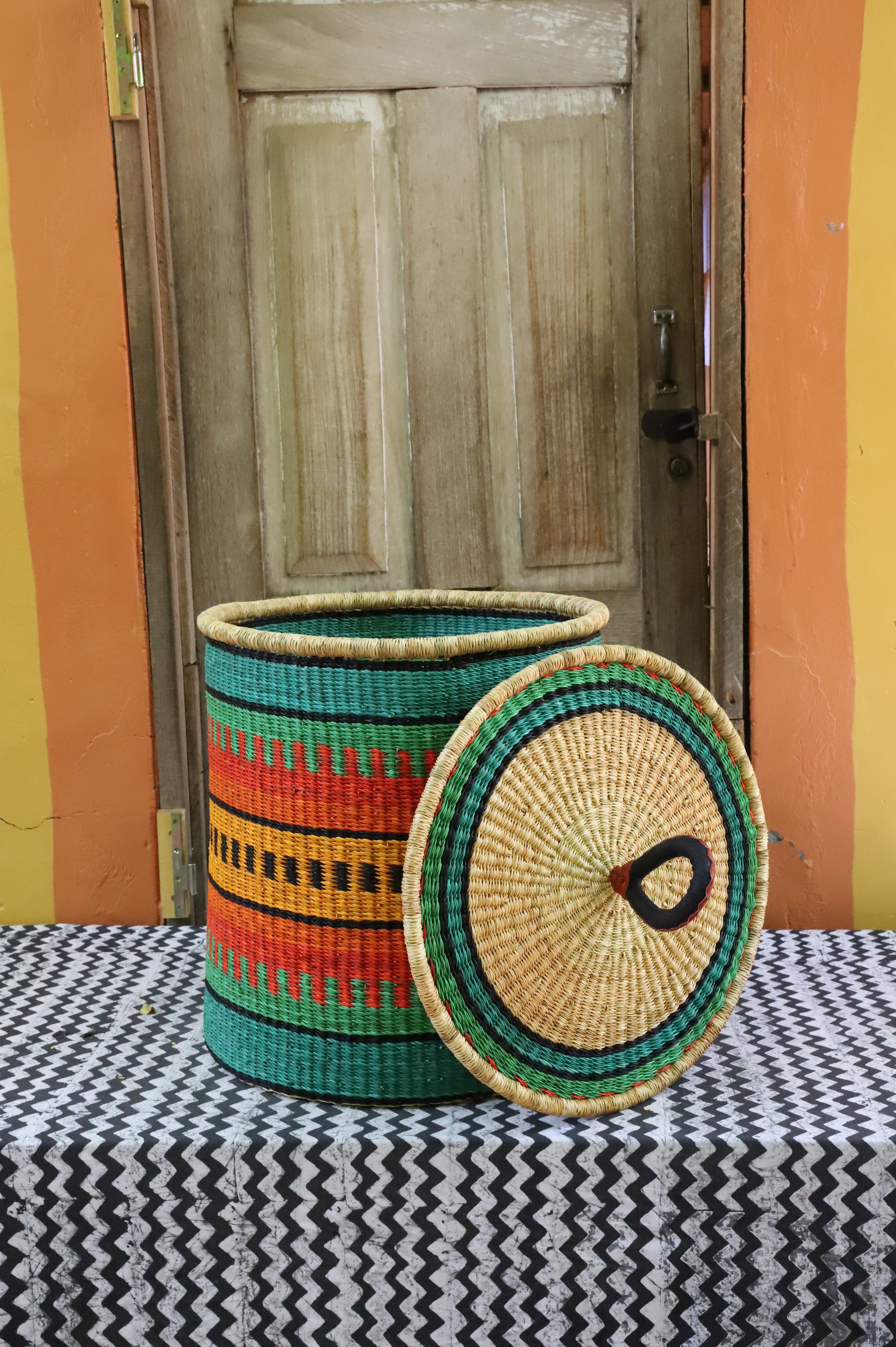 Large coiled-rim laundry basket in green, orange and black striped weave, lid resting alongside on a chevron cloth.