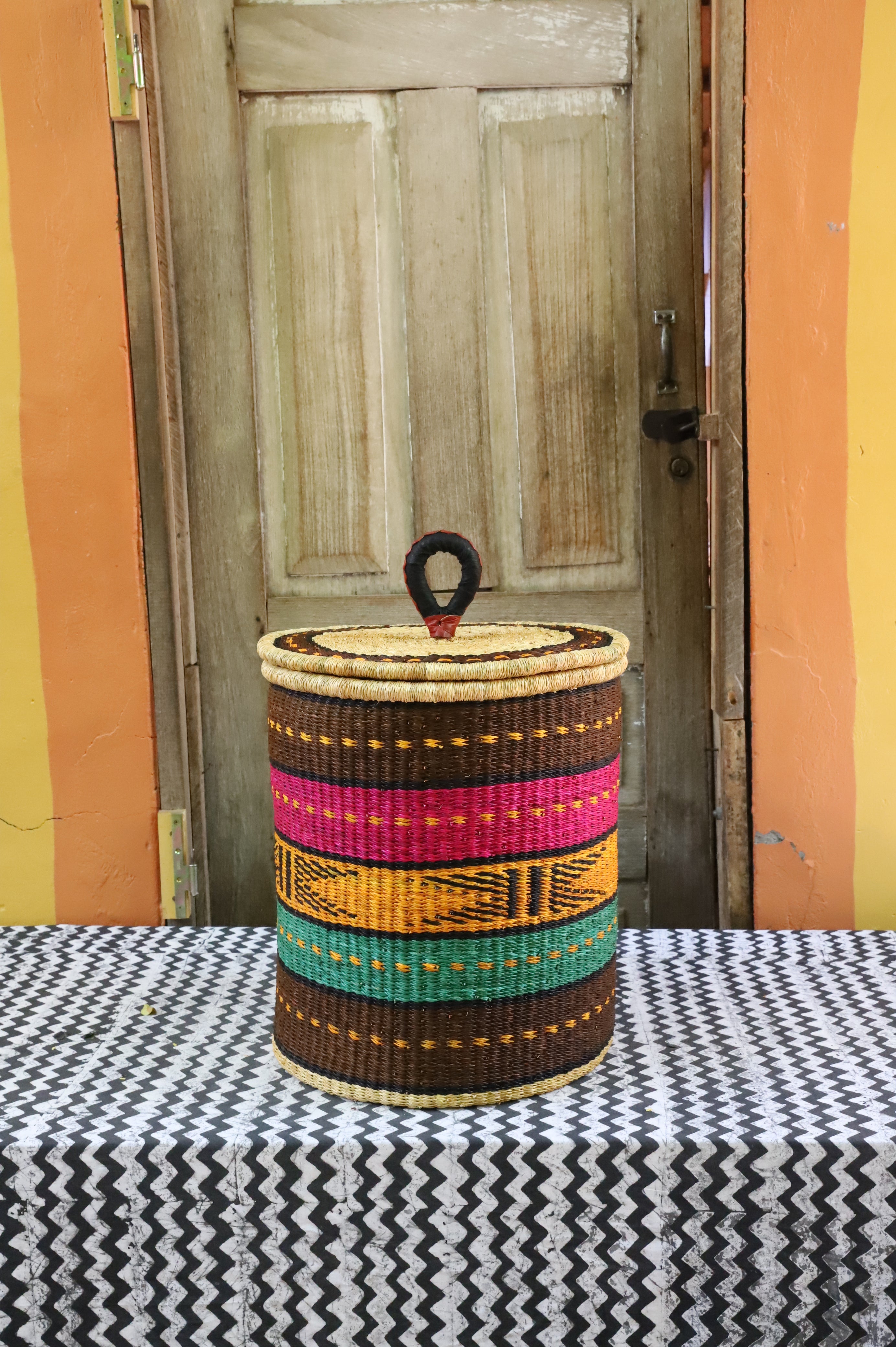 Coiled-rim lidded laundry basket in brown, pink, mustard and teal striped weave, styled on a chevron cloth.
