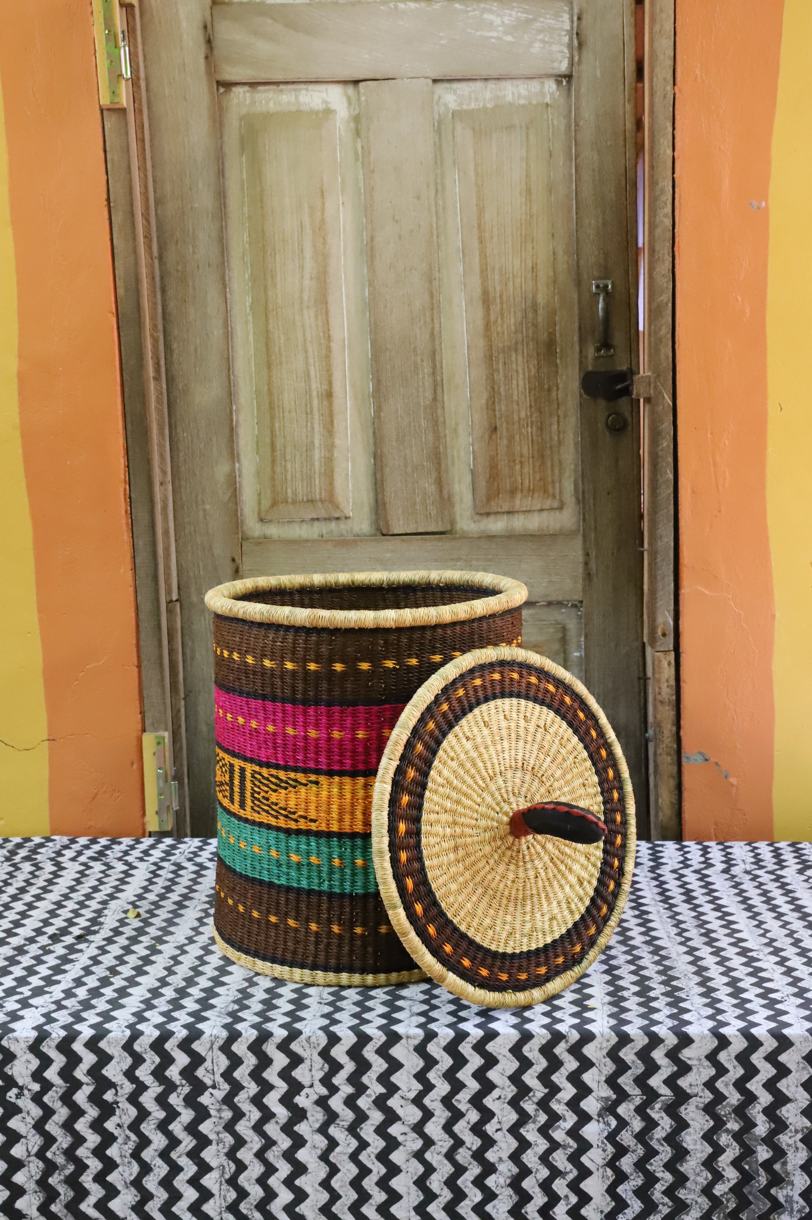 Coiled-rim laundry basket in brown, pink, mustard and teal striped weave, lid resting alongside on a chevron cloth.