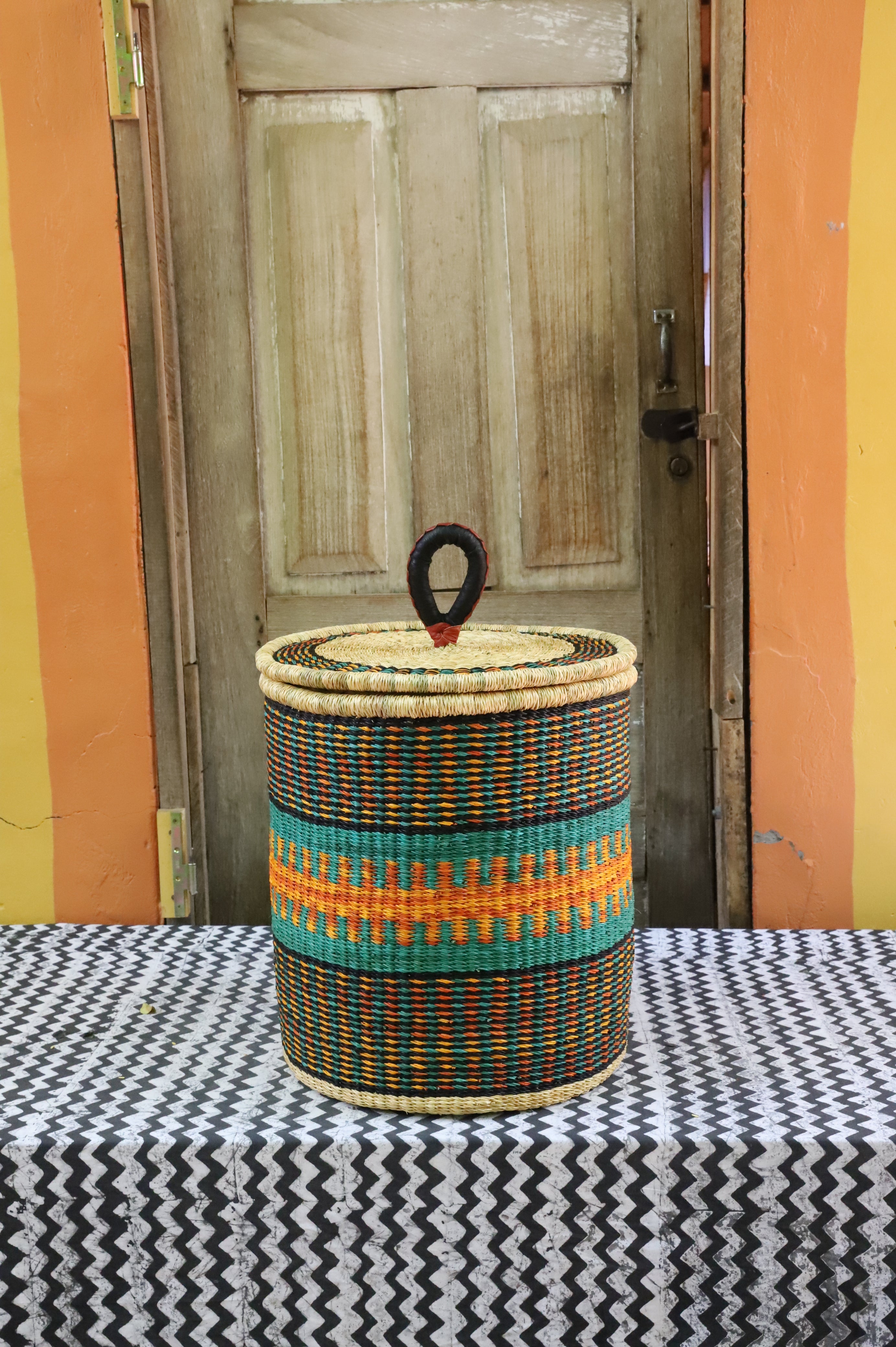 Coiled-rim lidded laundry basket in dark weave with turquoise and orange banding, styled on a chevron cloth.