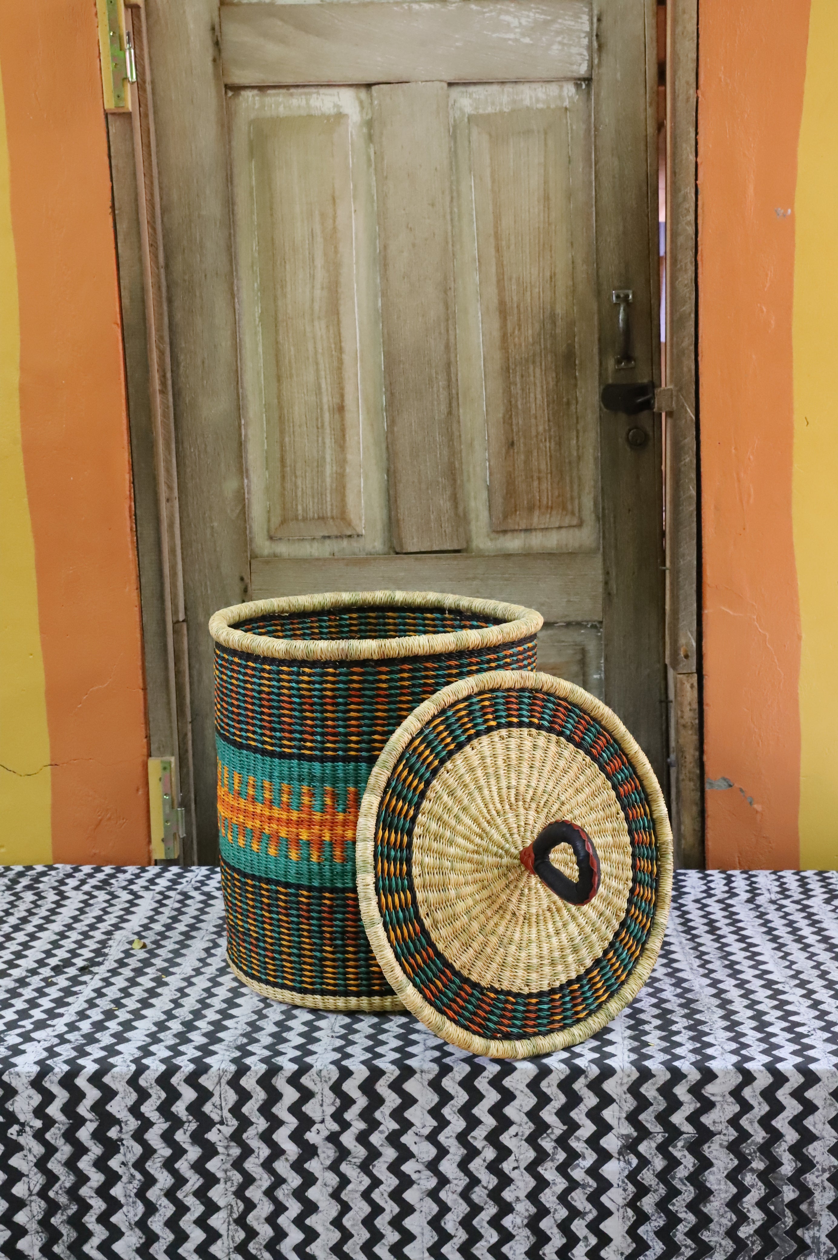 Coiled-rim laundry basket in dark weave with turquoise and orange banding, lid resting alongside on a chevron cloth.