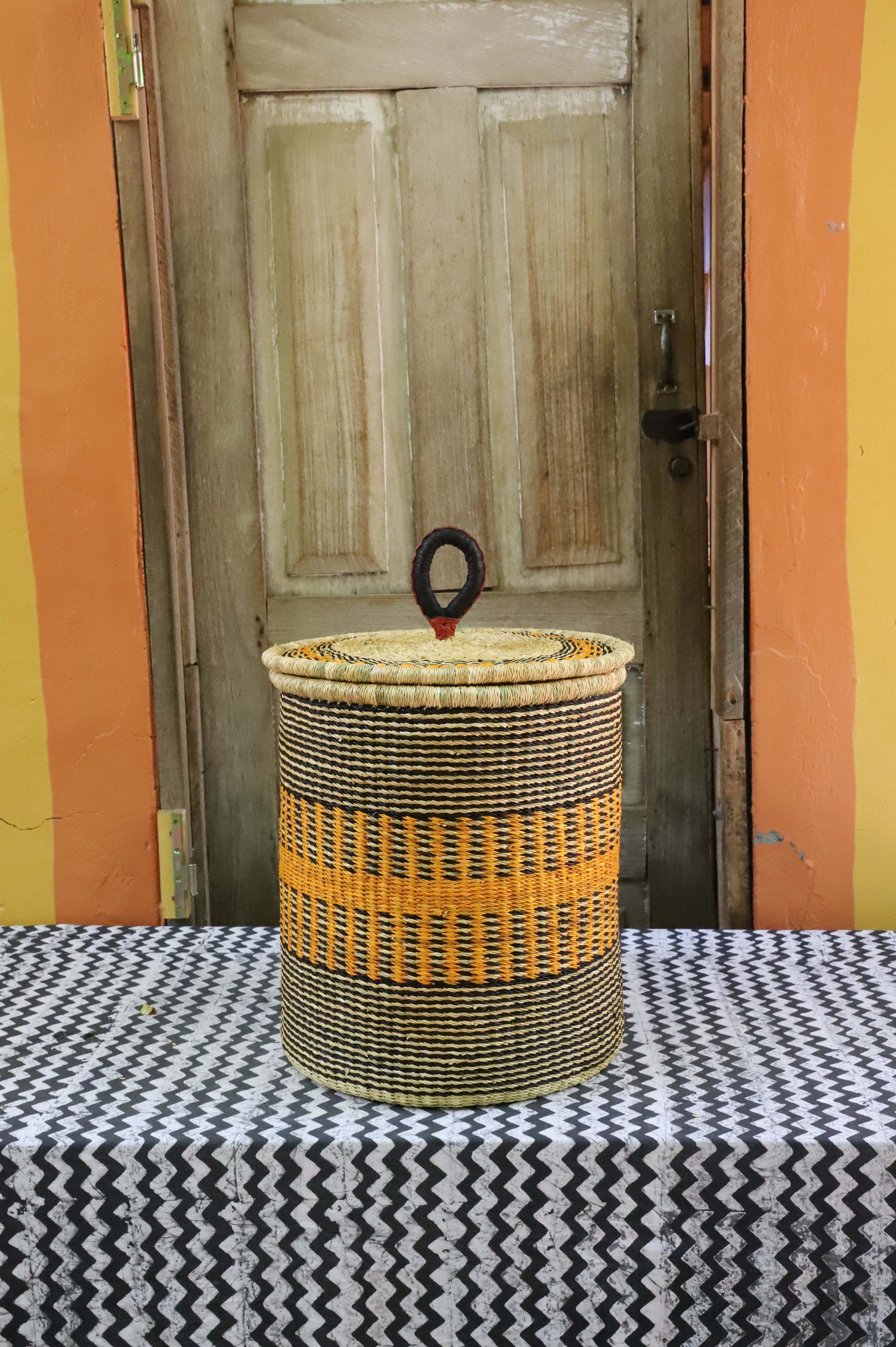 Coiled-rim lidded laundry basket in natural straw with a striped orange band, styled on a chevron cloth by a wooden door.