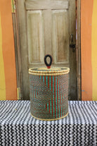 Coiled-rim lidded laundry basket in natural straw with teal and maroon banding, styled by a wooden door.