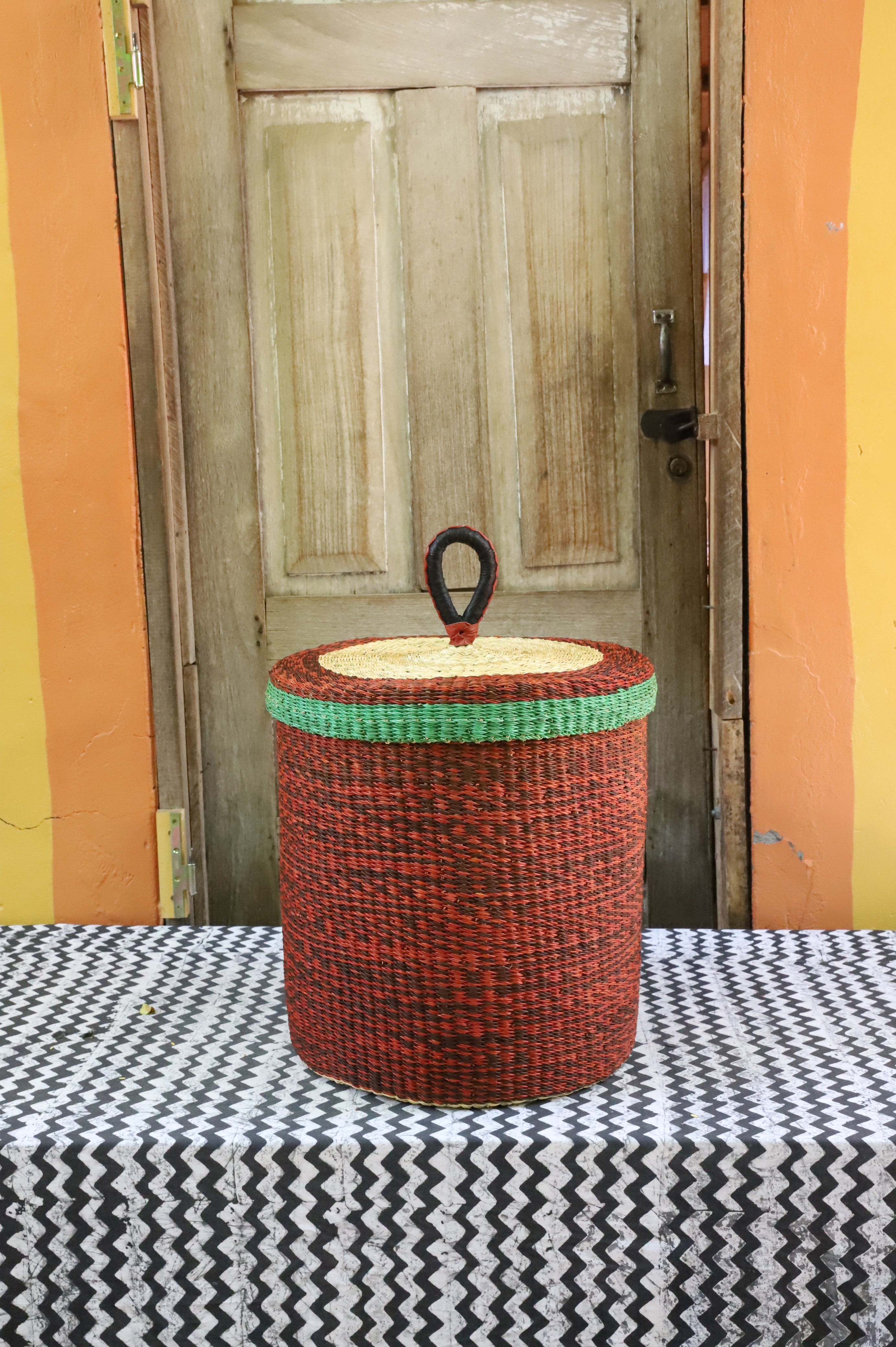 Lidded laundry basket in solid rust-red weave with a green rim trim, styled on a chevron cloth by a wooden door.