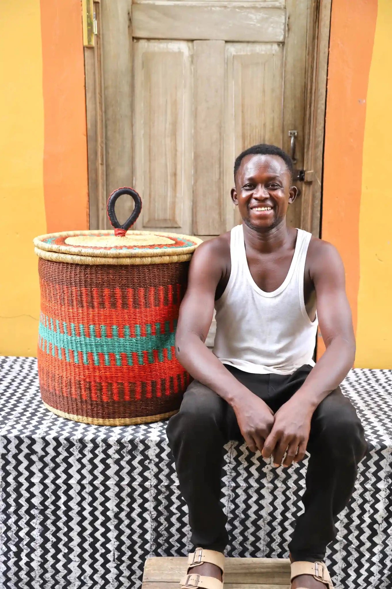Man sitting next to a woven basket against a wooden  door.