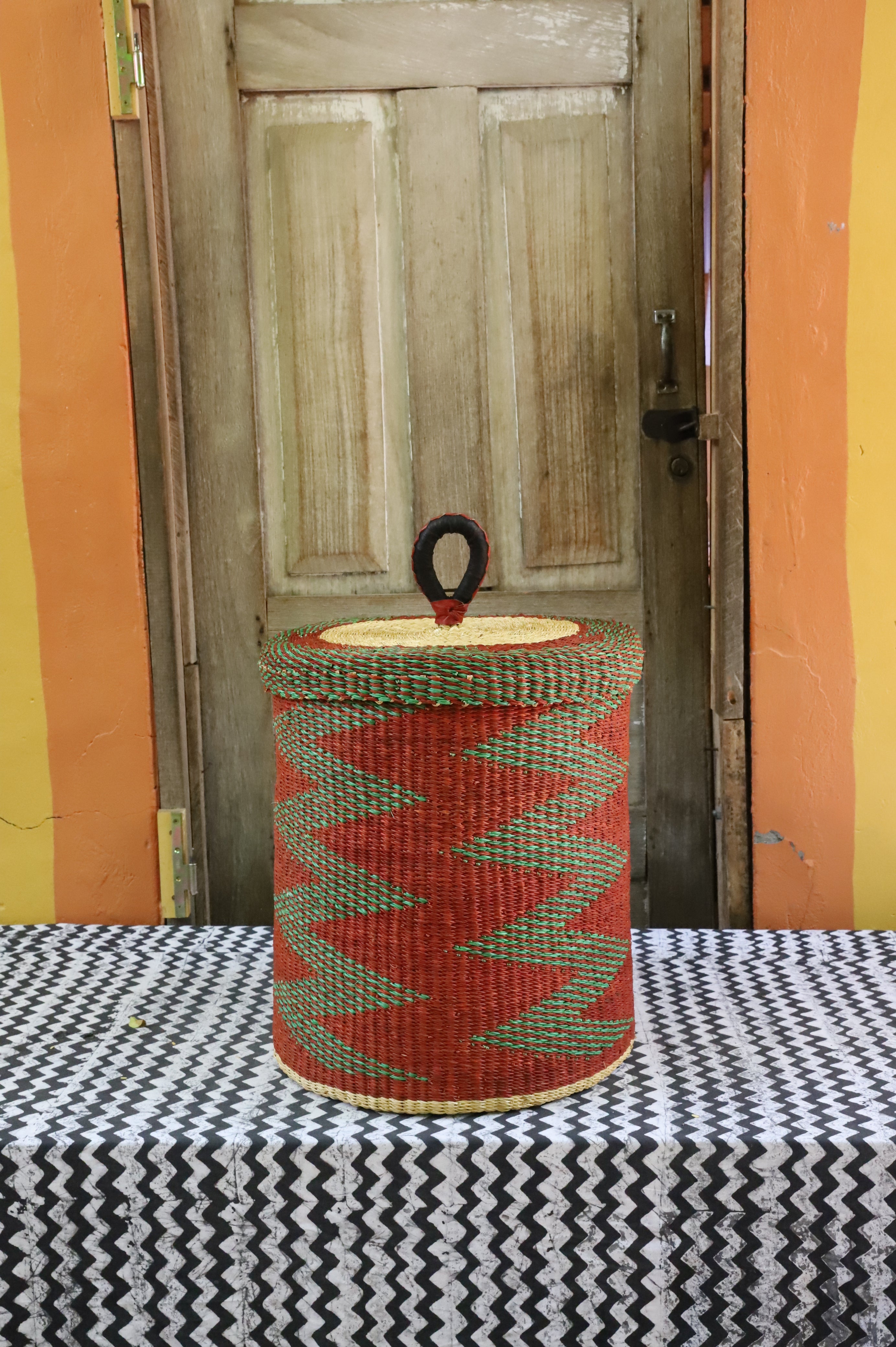 Lidded laundry basket in rust and green zigzag weave, styled on a chevron cloth by a wooden door.