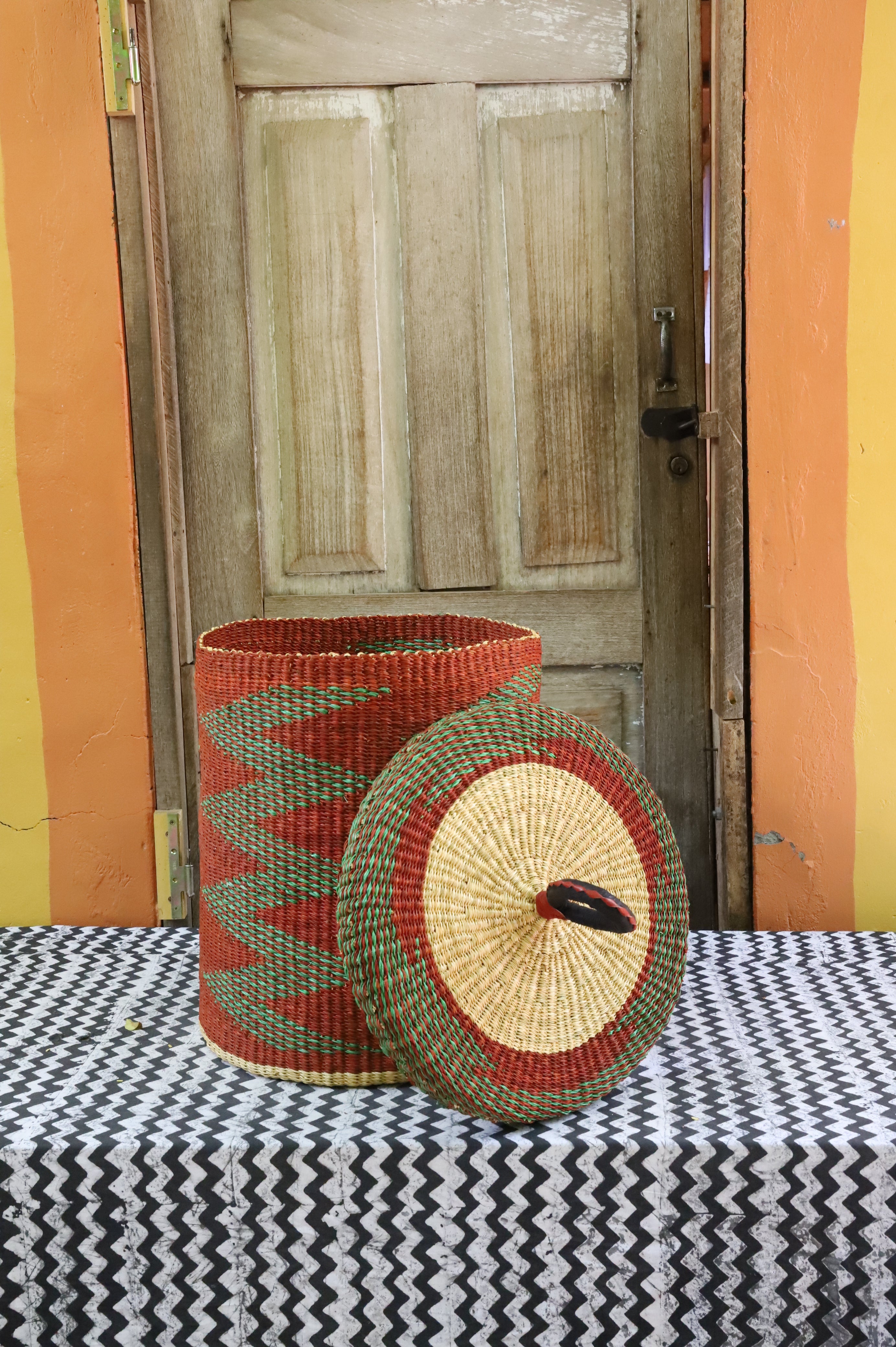 Lidded laundry basket in rust and green zigzag weave, lid resting alongside showing the straw underside.