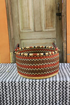 Handwoven Bolga basket in natural straw with maroon and black banded weave, styled on a chevron cloth by a wooden door.