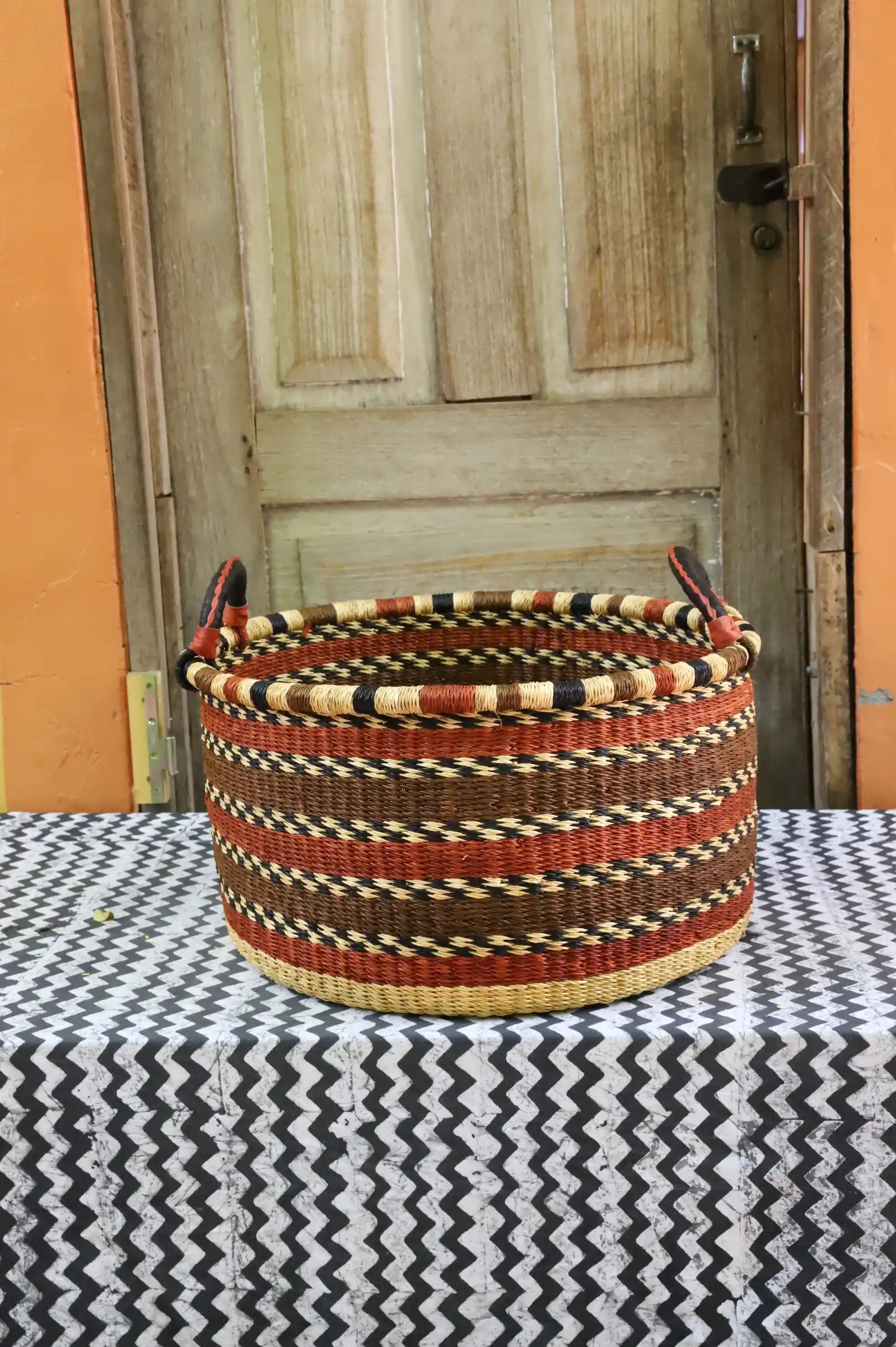 Handwoven Bolga basket in natural straw with maroon and black banded weave, styled on a chevron cloth by a wooden door.