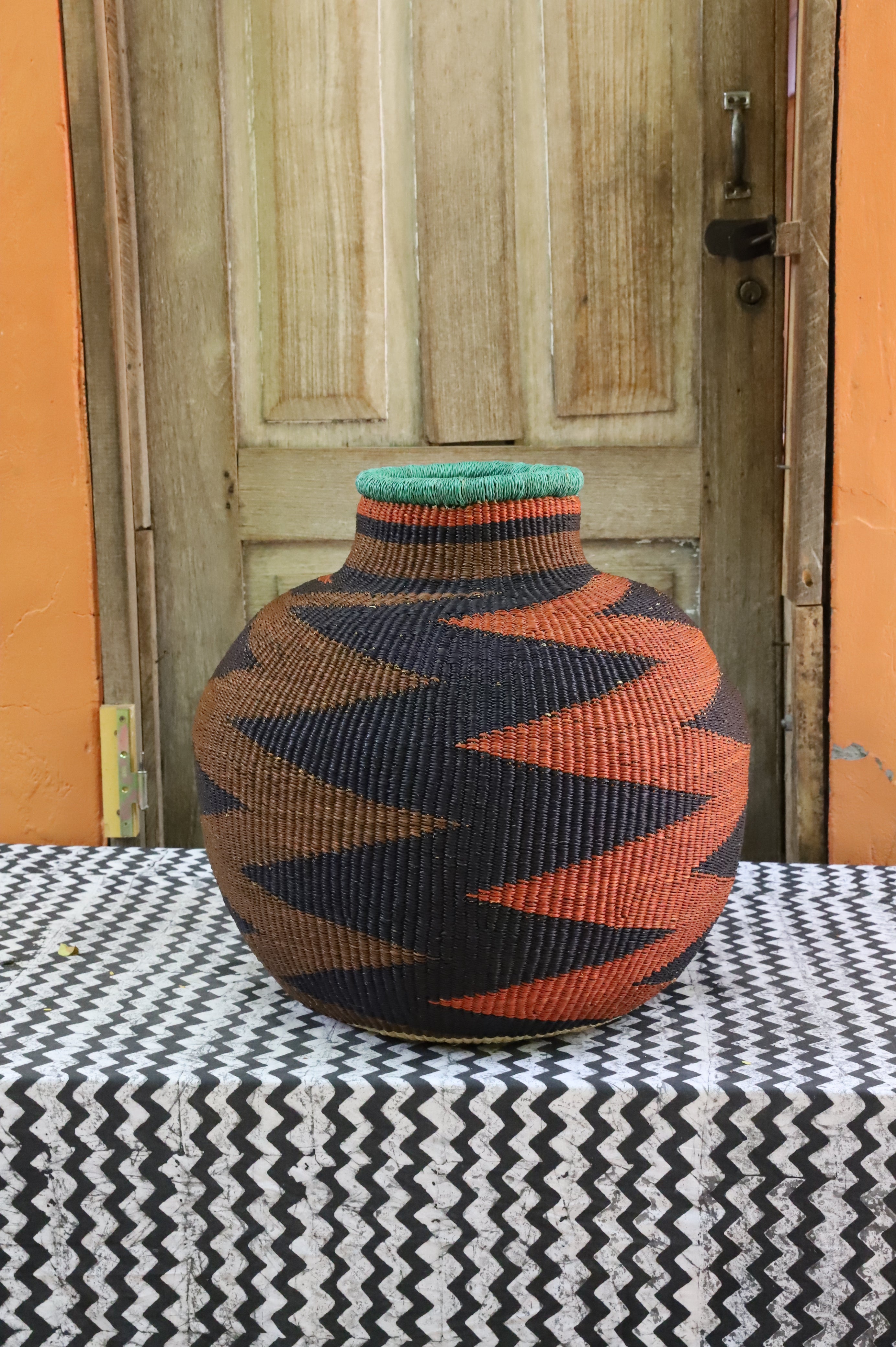 Bulbous 10 Cows décor basket in black and rust zigzag weave with a teal rim, styled on a chevron cloth by a wooden door.