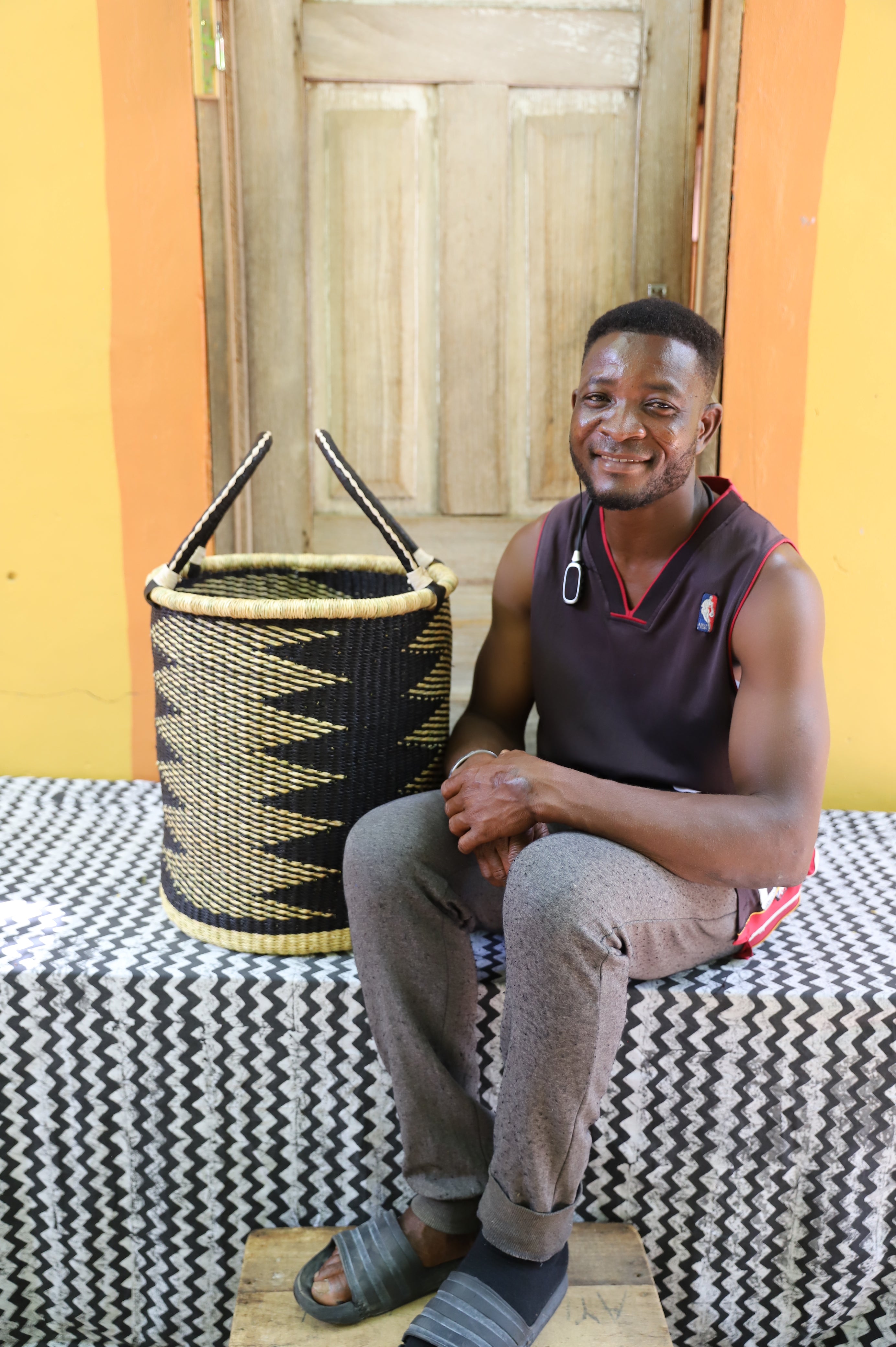 Open laundry basket in black and natural straw zigzag weave, shown beside a seated person for scale.