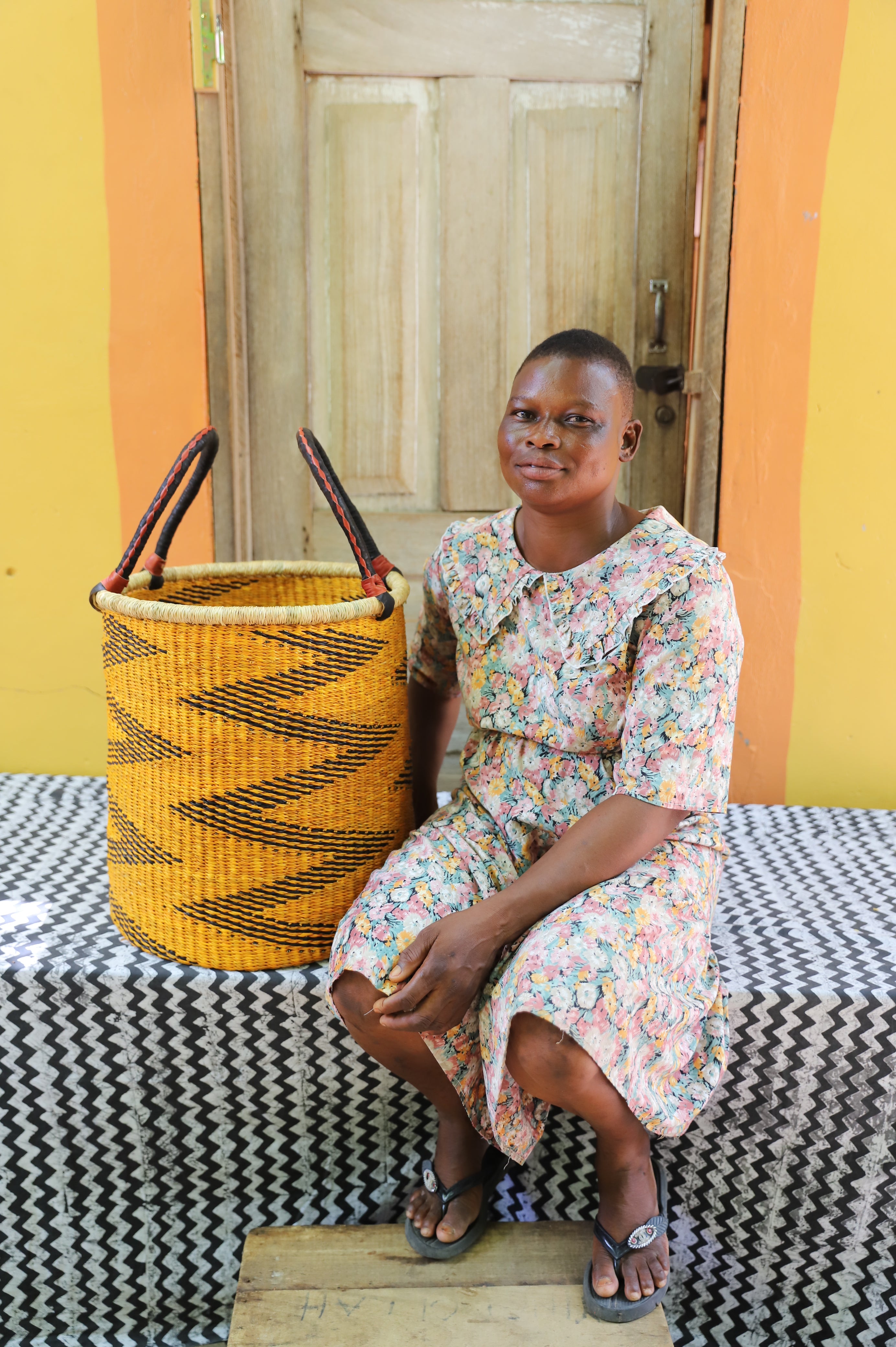 Open laundry basket in mustard and black zigzag weave with arched handles, shown beside a seated person for scale.