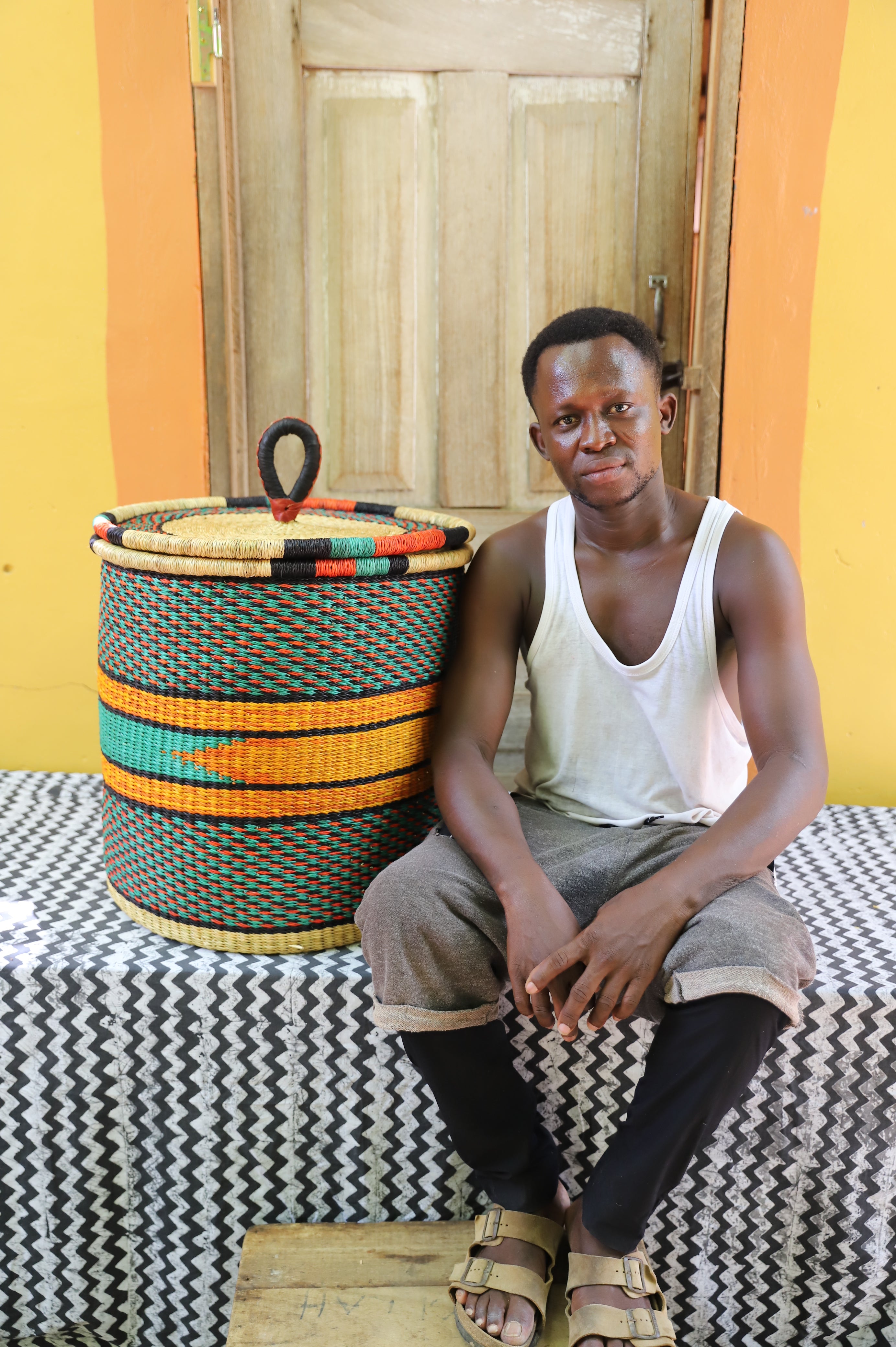 Large coiled-rim lidded laundry basket in dark weave with orange and black banding, shown beside a seated person for scale.