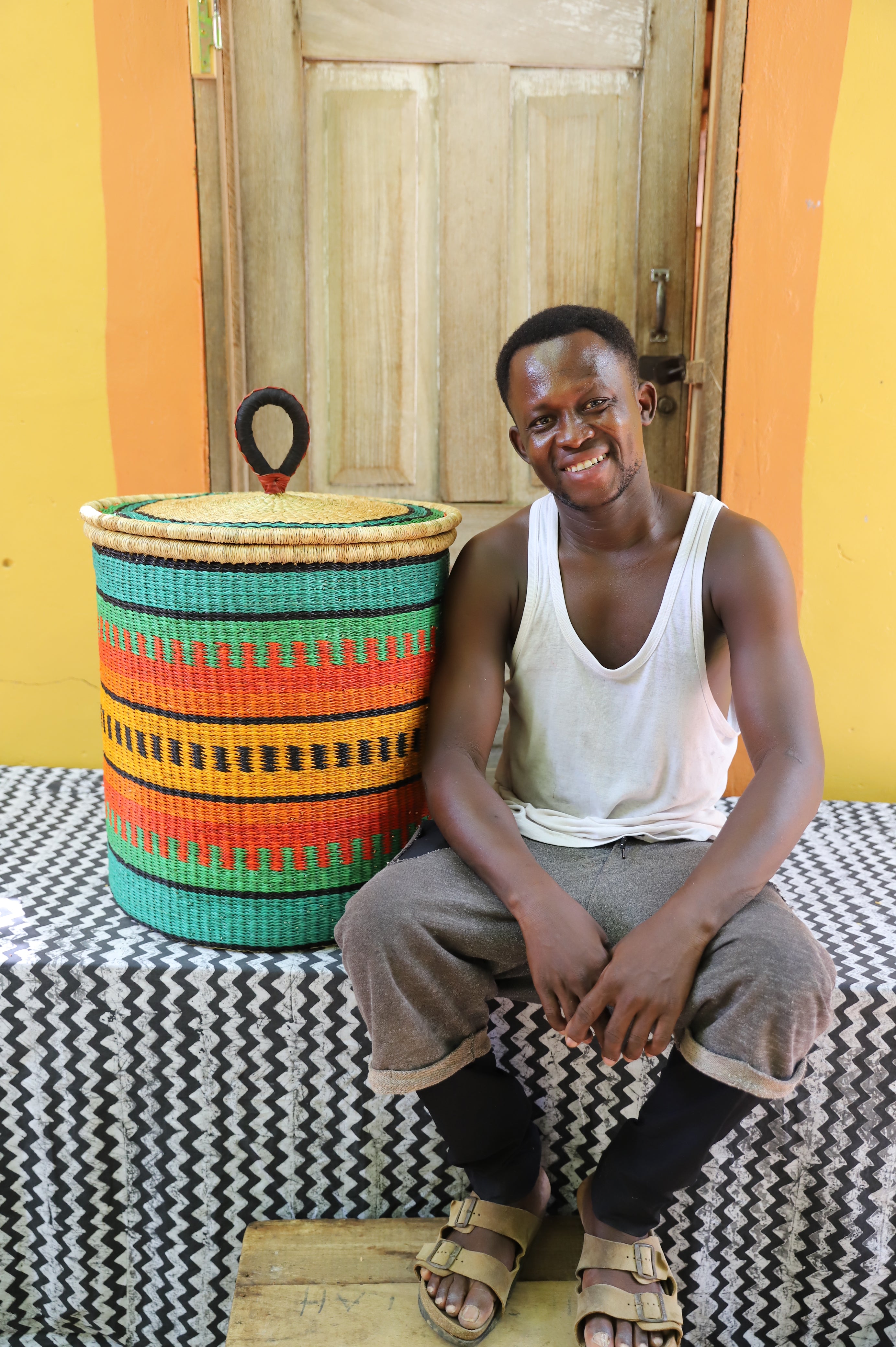 Large coiled-rim lidded laundry basket in green, orange and black striped weave, shown beside a seated person for scale.