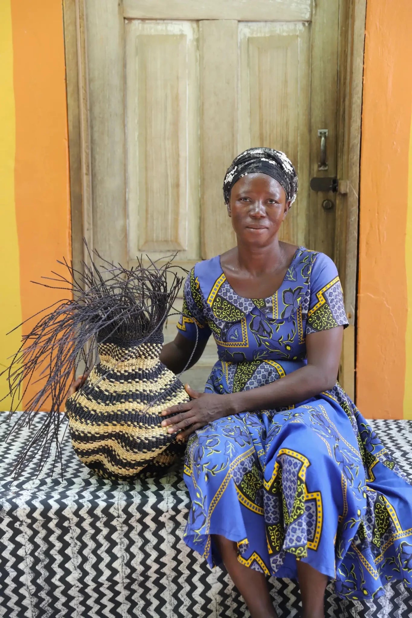 A women sitting next to a colorful, handwoven piece sitting on a black and white cloth with decorative straw branching out. 