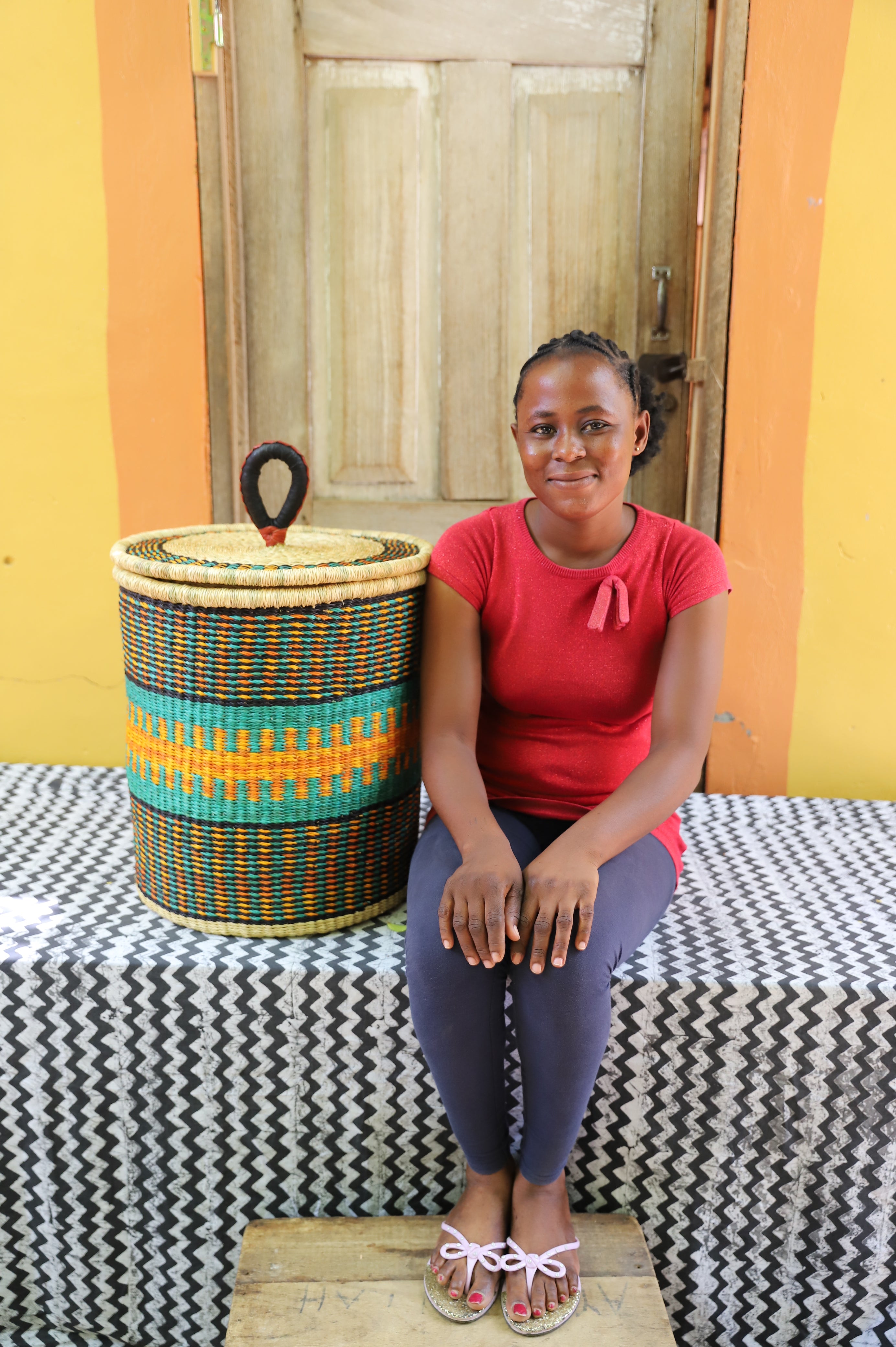 Coiled-rim lidded laundry basket in dark weave with turquoise and orange banding, shown beside a seated person for scale.