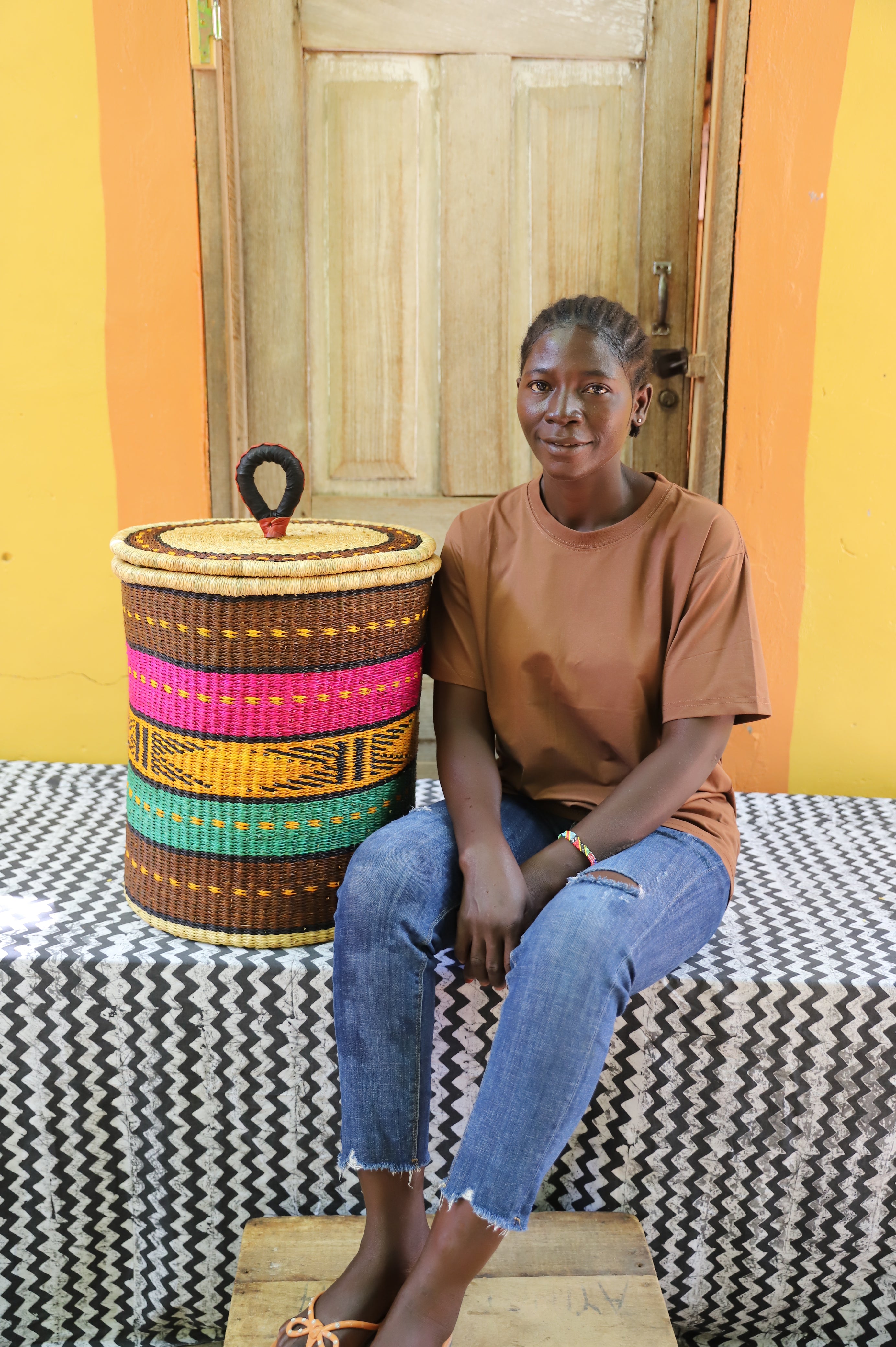 Coiled-rim lidded laundry basket in brown, pink, mustard and teal striped weave, shown beside a seated person for scale.