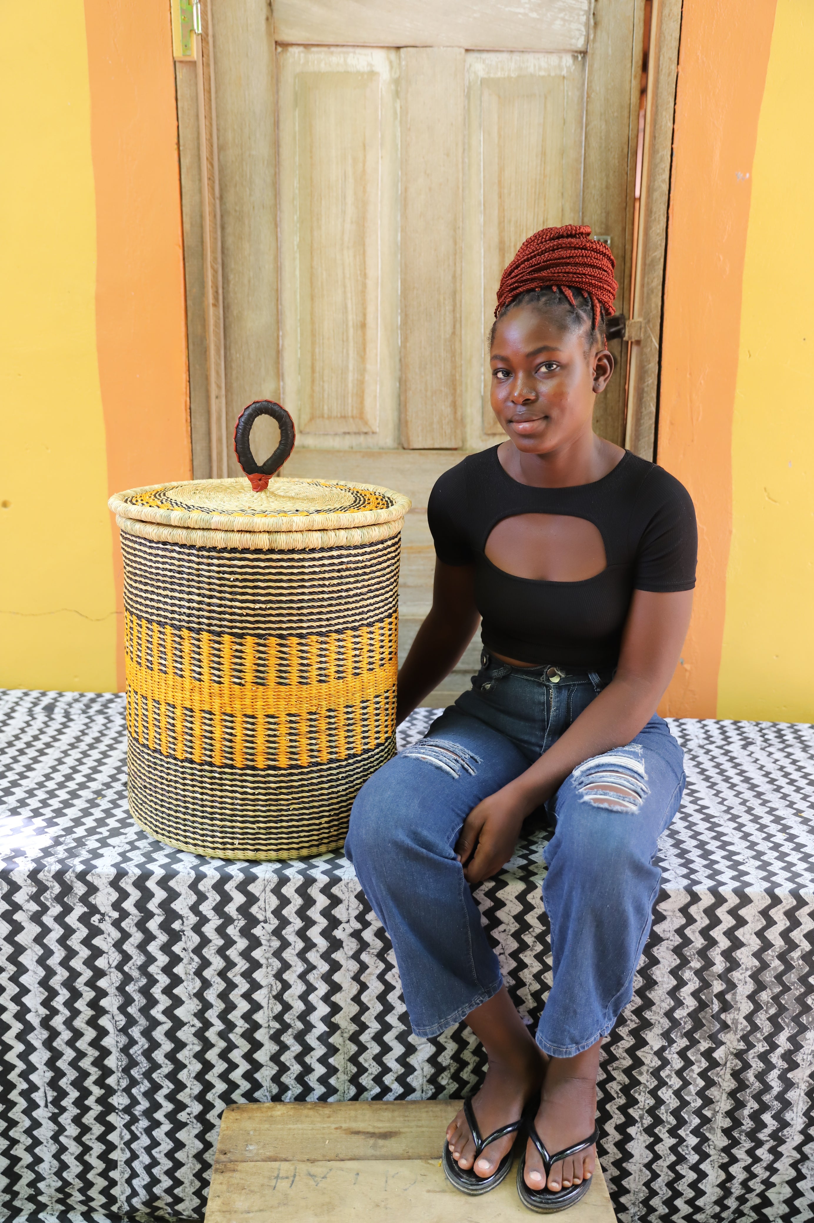 Coiled-rim lidded laundry basket in natural straw with a striped orange band, shown beside a seated person for scale.