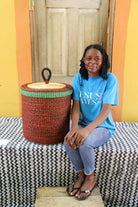 Lidded laundry basket in solid rust-red weave, shown beside a seated person for scale.