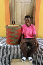 Lidded laundry basket in rust and green zigzag weave, shown beside a seated person for scale.