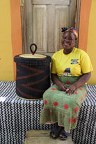 Lidded laundry basket in dark brown elephant grass, shown beside a seated person for scale.
