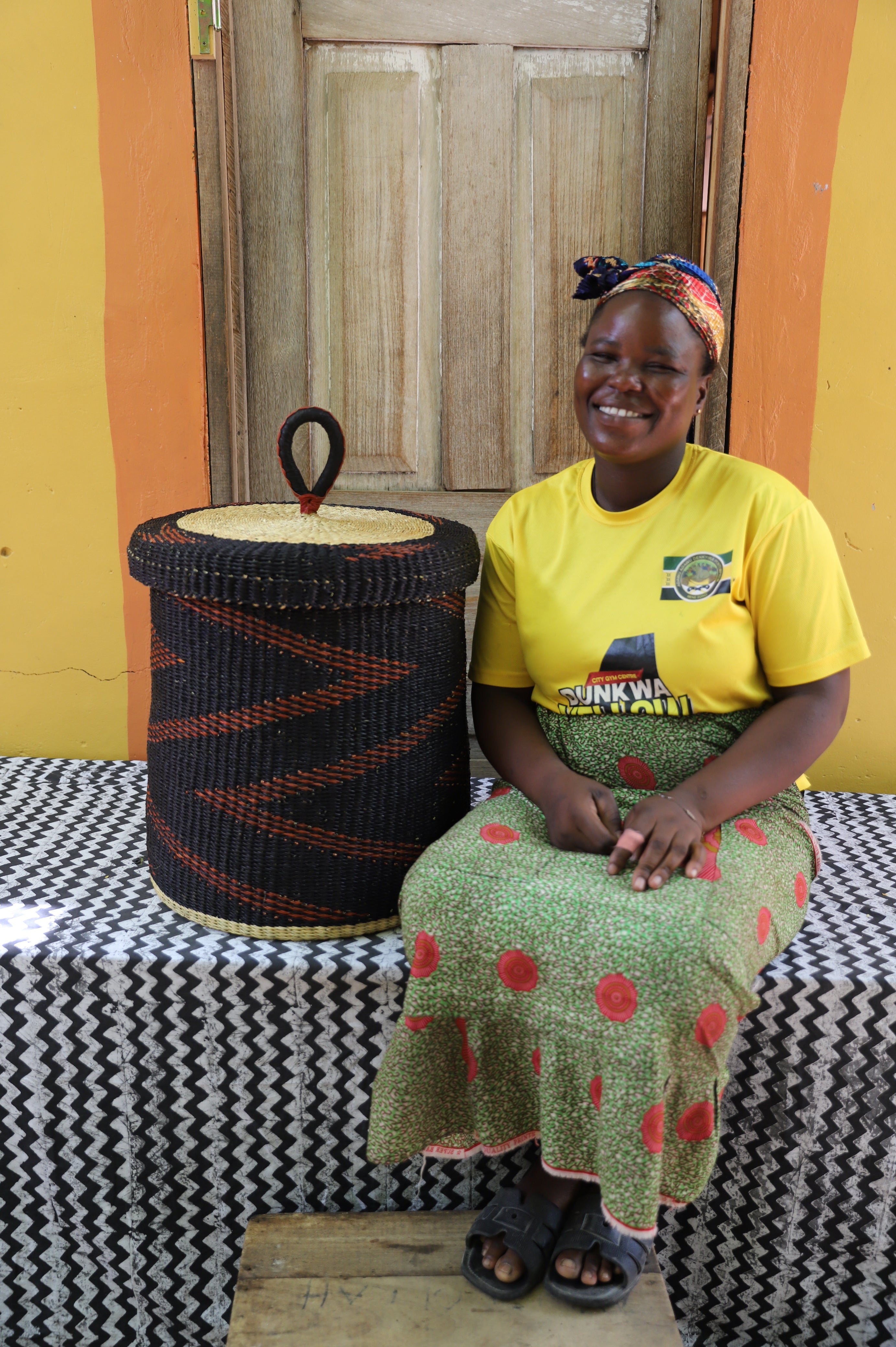 Lidded laundry basket in dark brown elephant grass, shown beside a seated person for scale.