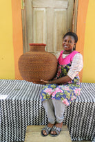 Bulbous décor basket in natural straw with a rust-dyed neck band, held by a seated person for scale.