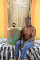 Coiled-rim lidded laundry basket in natural straw with teal and maroon banding, shown beside a seated person for scale.
