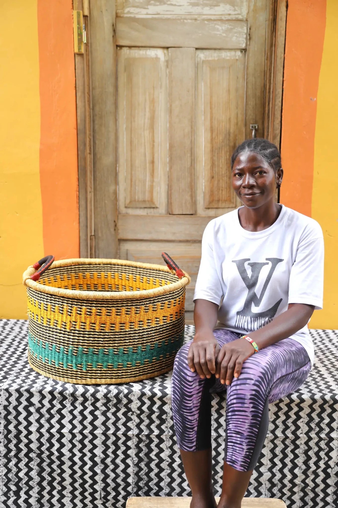 Woven Tub (Large) with woman weaver handwoven in Ghana sitting on black and white cloth and wooden door in background
