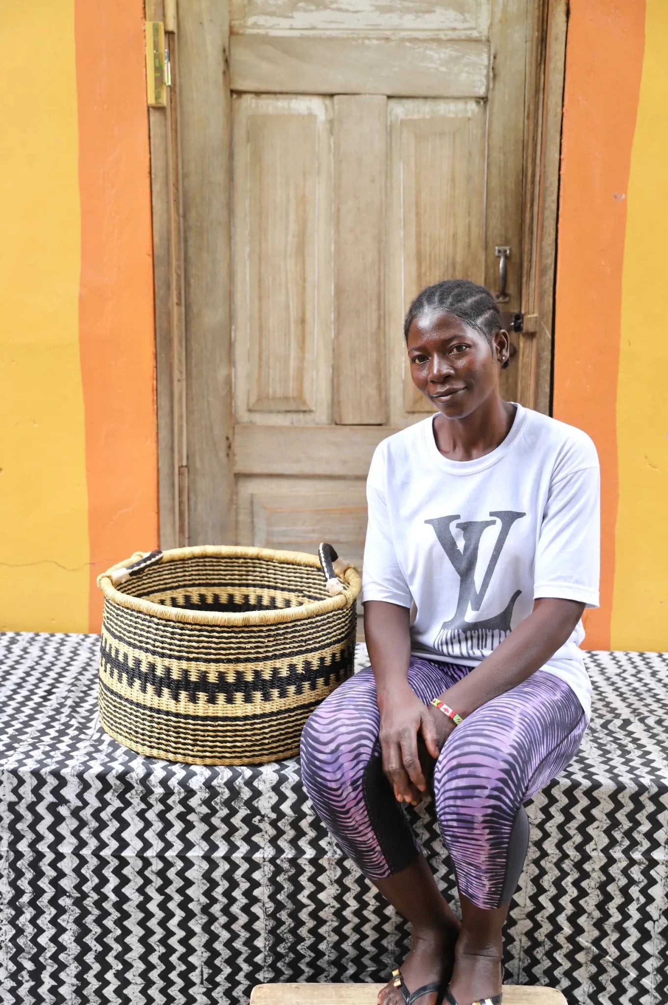 Woven Tub (Small) with woman weaver handwoven in Ghana sitting on black and white cloth and wooden door in background

