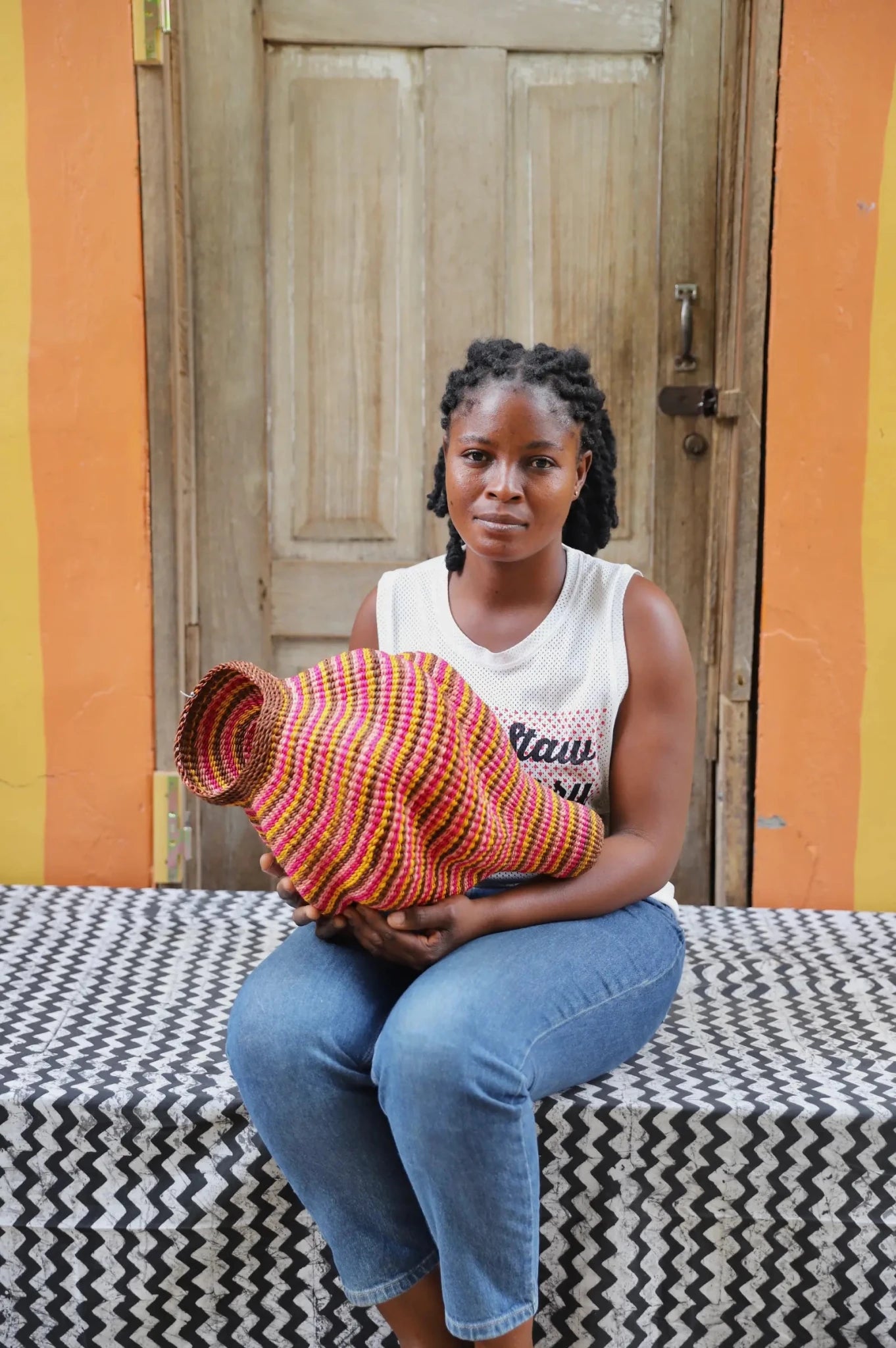 Woman holding a woven conch piece on a black and white patterned surface with a wooden door in the background.
