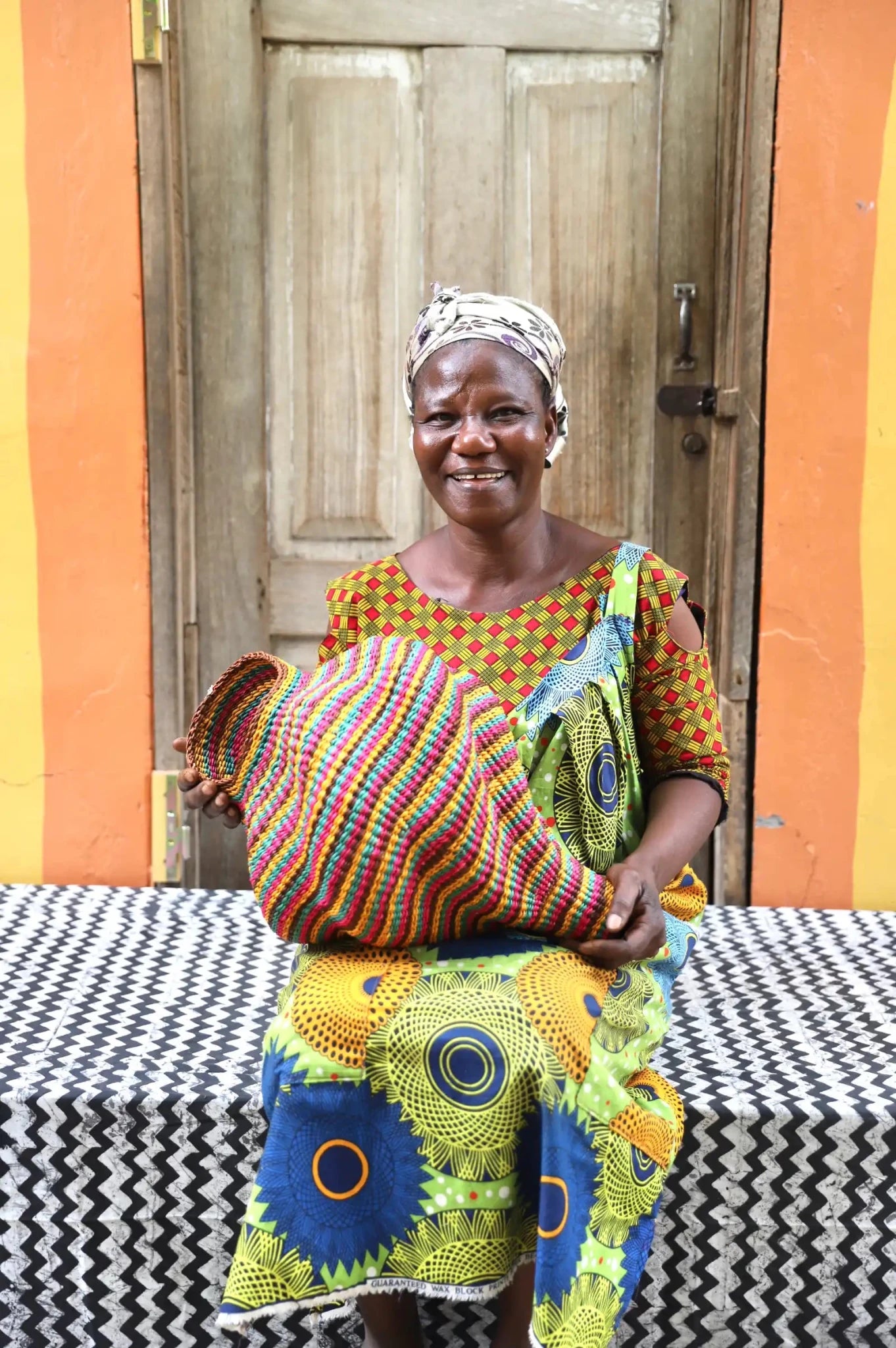 Woman sitting with a woven conch piece on a black and white patterned surface with a wooden door in the background.
