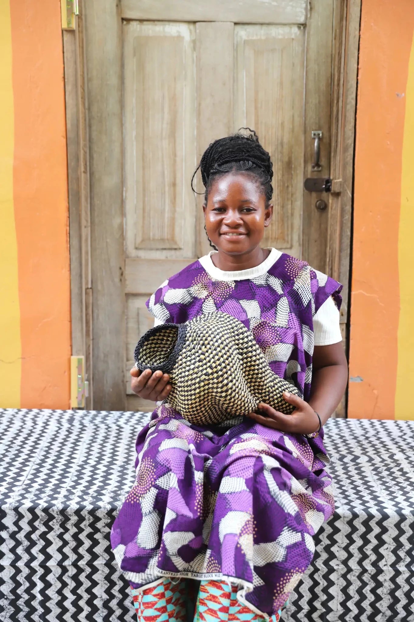 Woman holding a woven conch piece on a black and white patterned surface with a wooden door in the background.