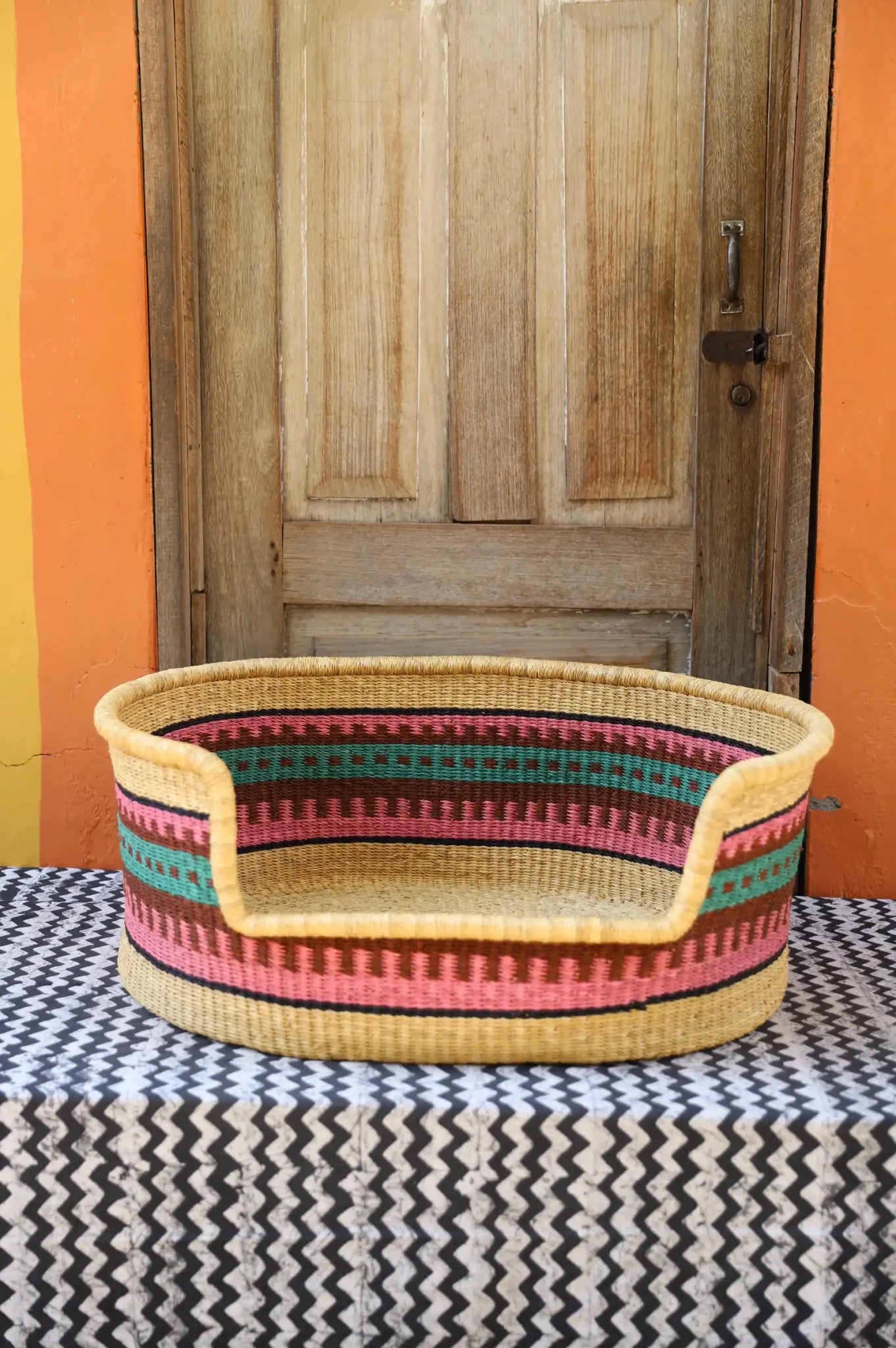 Woven pet bed with geometric pattern on a textured surface in front of a wooden door.
