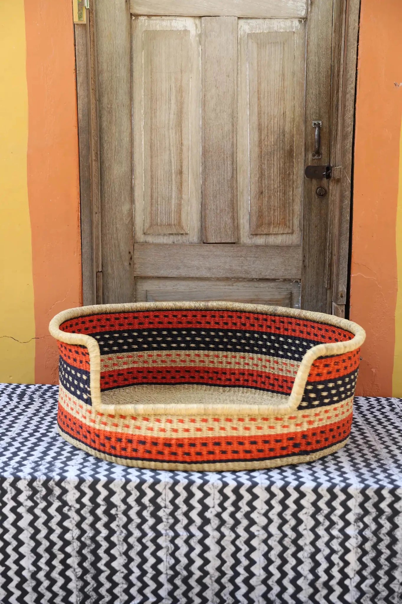 Woven pet bed with geometric pattern on a textured surface in front of a wooden door.