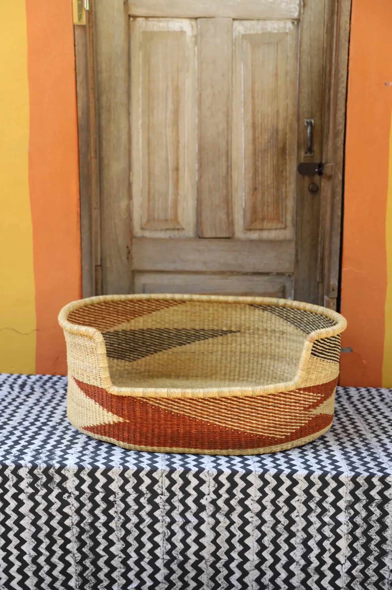 Woven pet bed with geometric pattern on a textured surface in front of a wooden door.