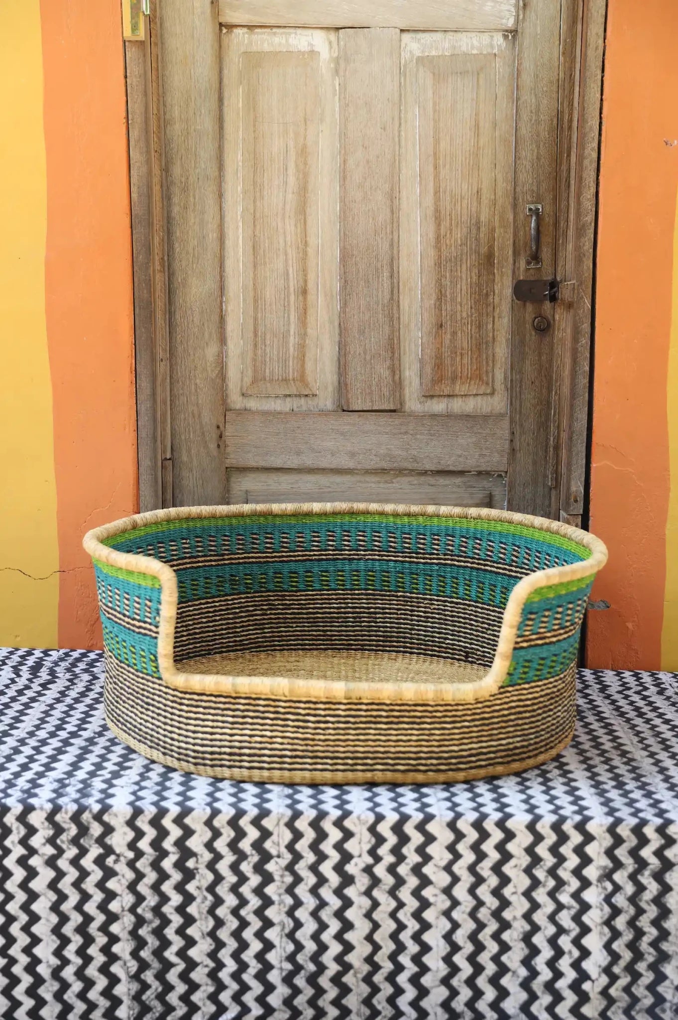 Woven pet bed with geometric pattern on a textured surface in front of a wooden door.