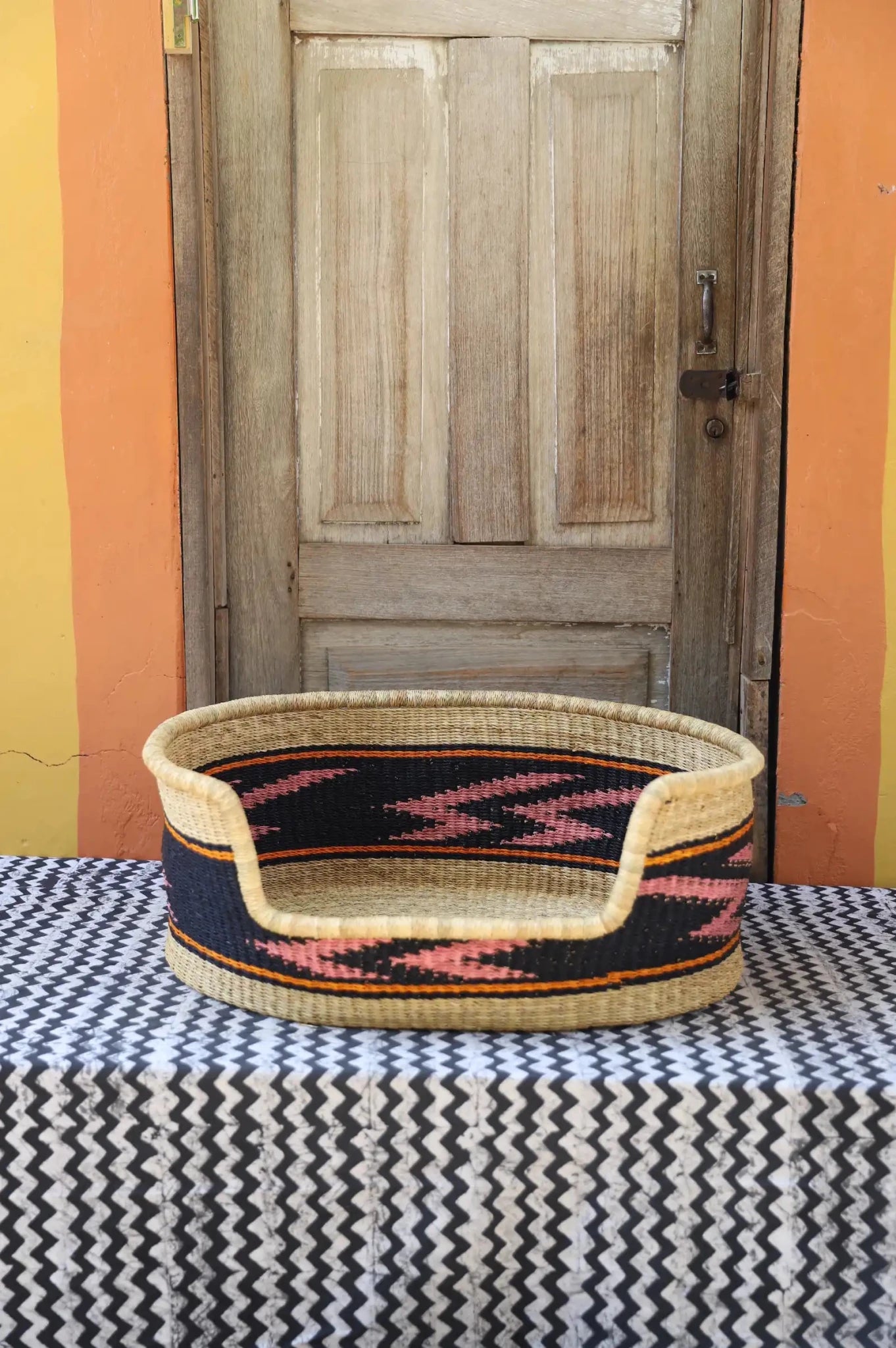 Woven pet bed with geometric pattern on a textured surface in front of a wooden door.