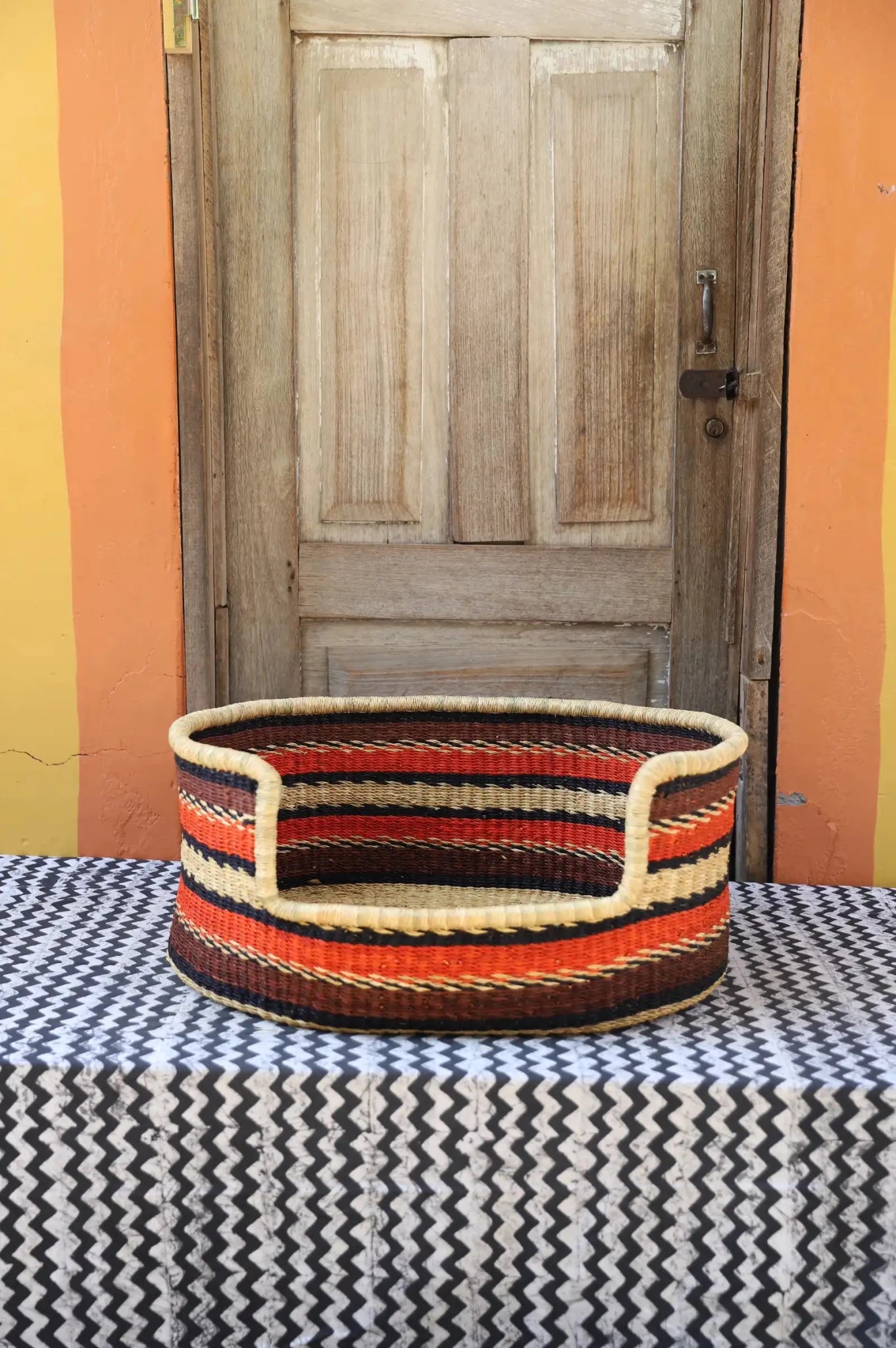 Woven pet bed with geometric pattern on a textured surface in front of a wooden door.