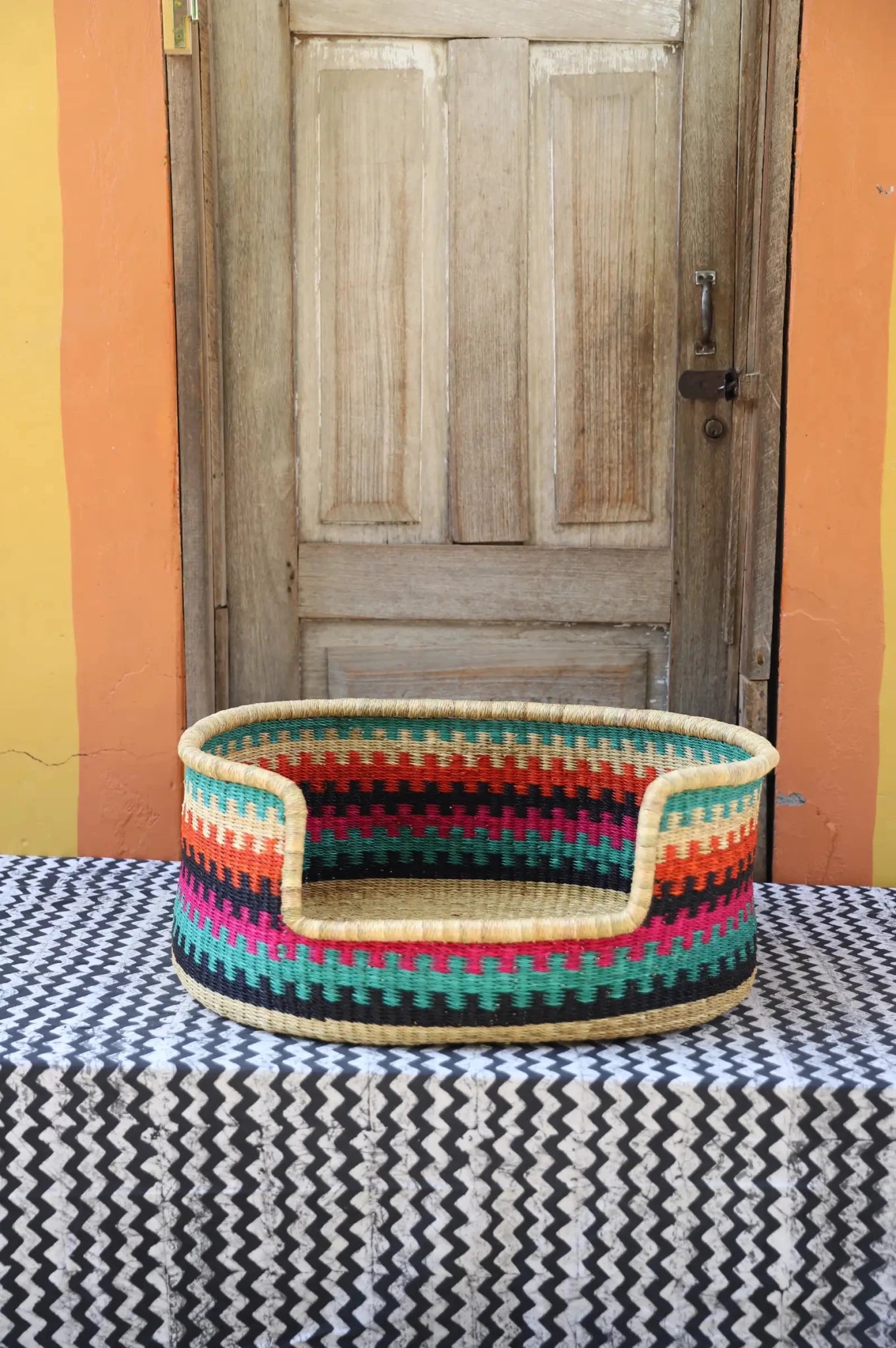 Woven pet bed with geometric pattern on a textured surface in front of a wooden door.