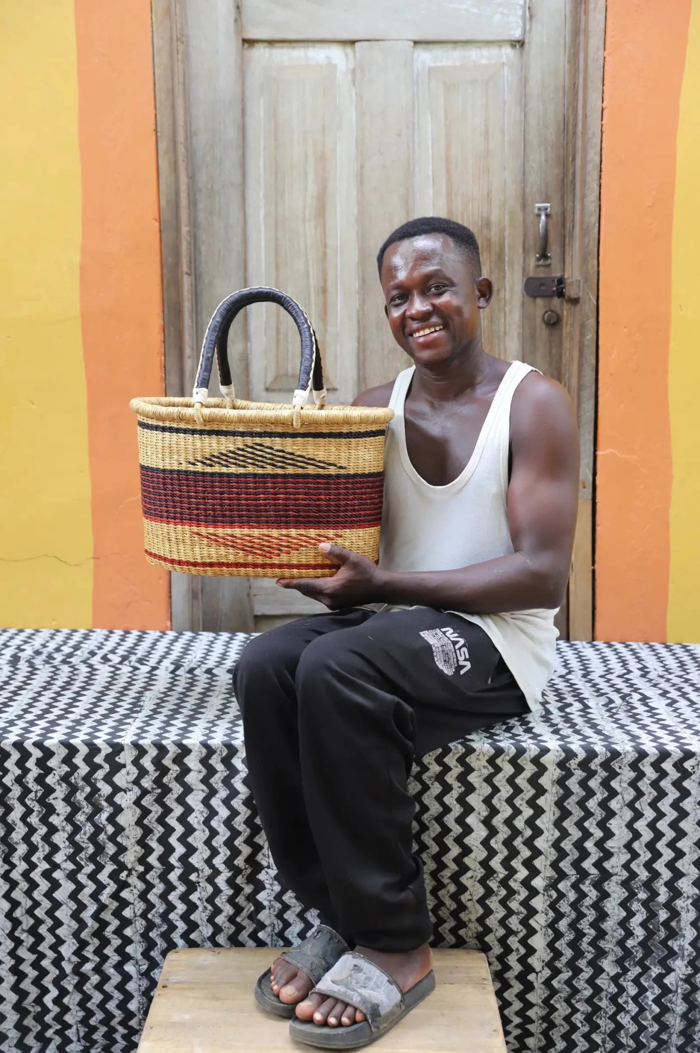 Man seated holding a large woven carry basket with bold burgundy, red and yellow stripe bands and a dark leather handle