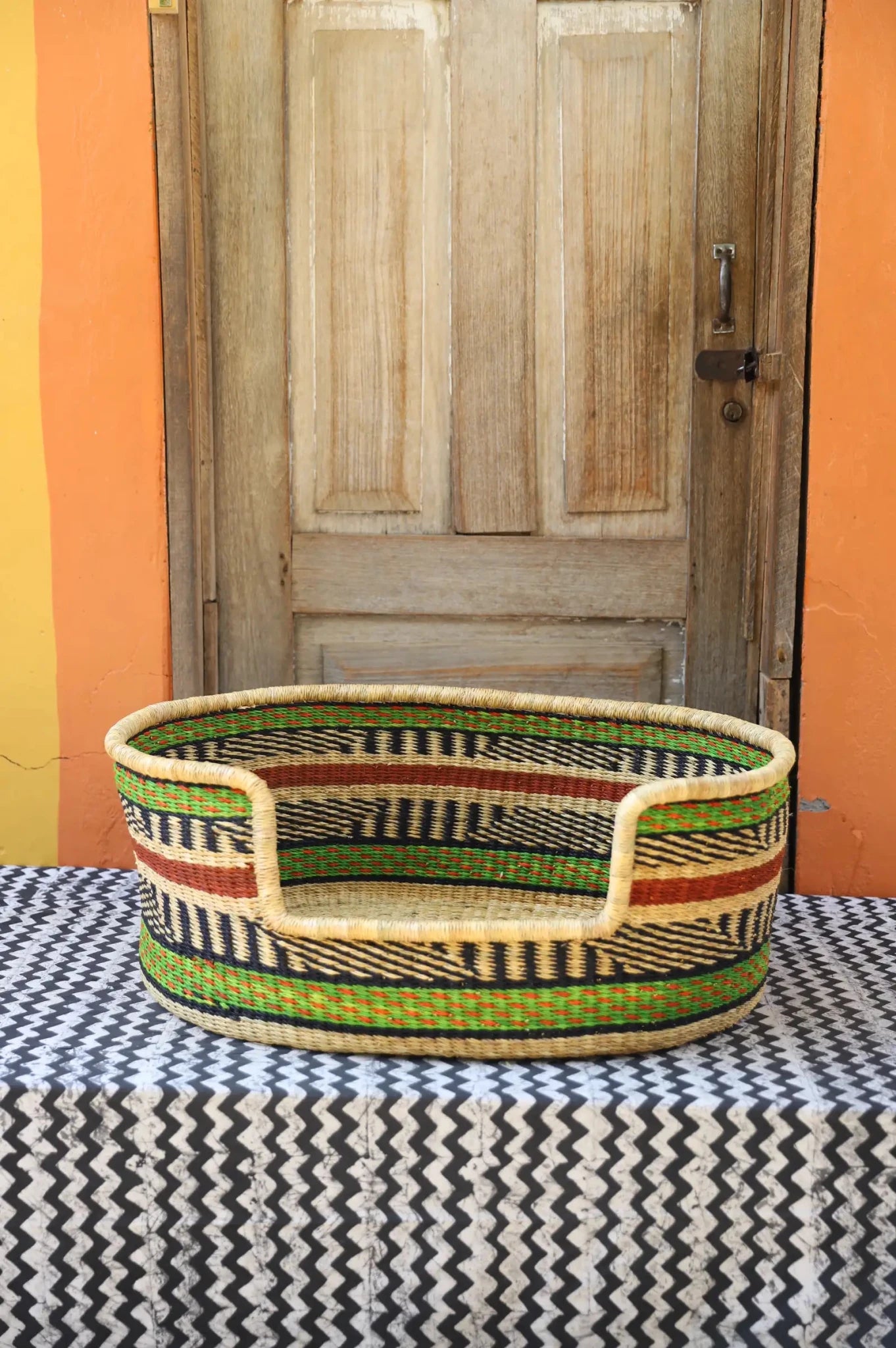 Woven pet bed with geometric pattern on a textured surface in front of a wooden door.
