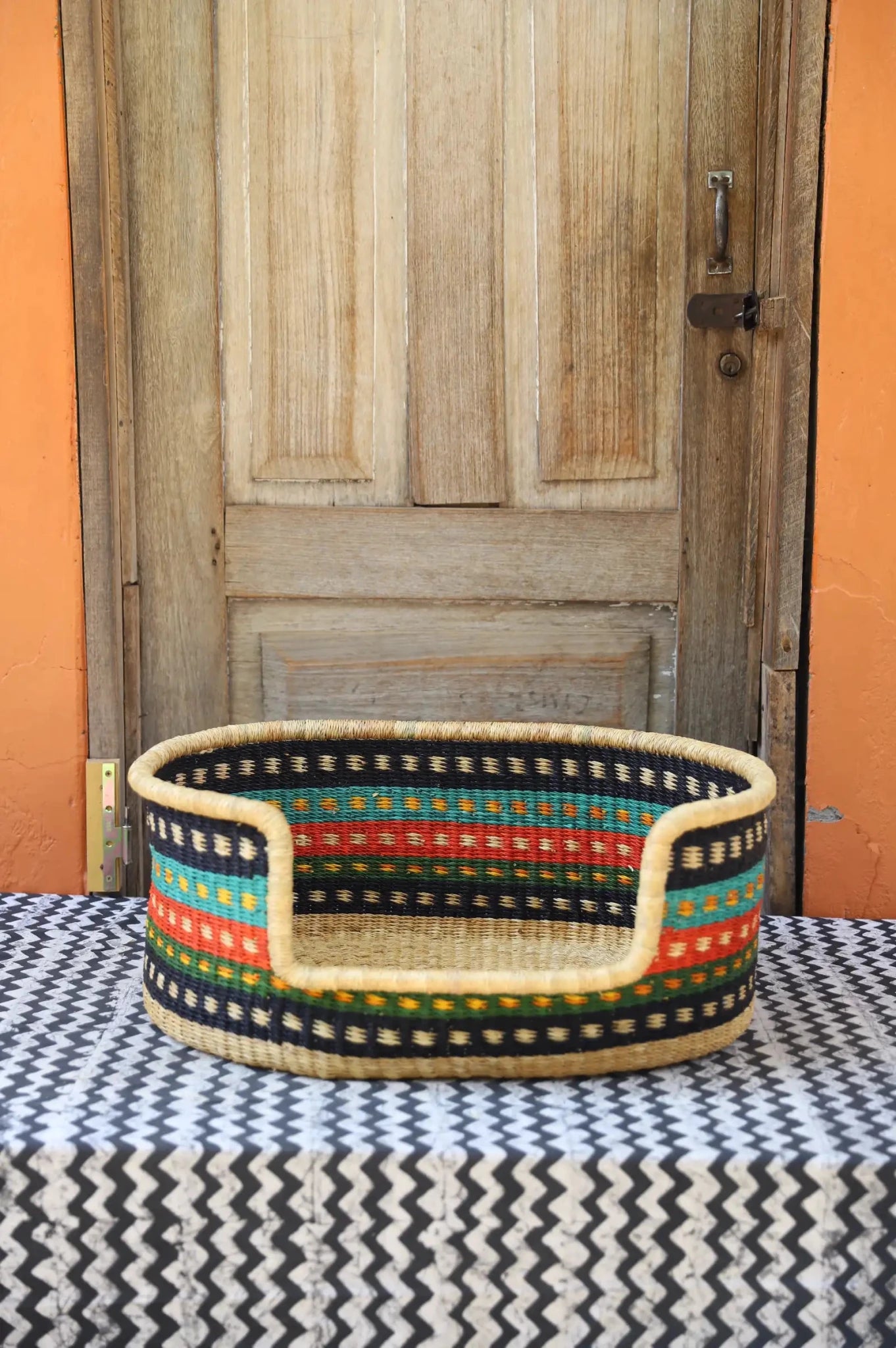Woven pet bed with geometric pattern on a textured surface in front of a wooden door.