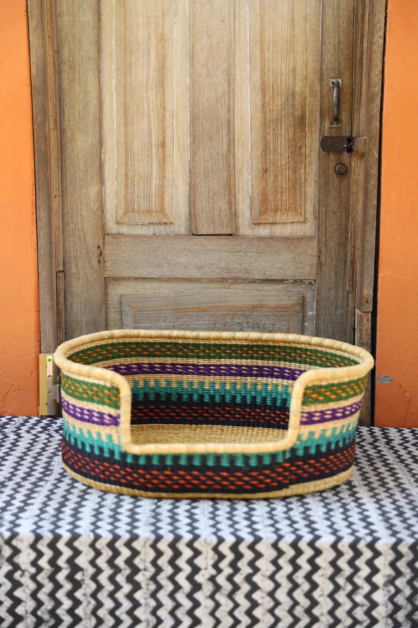 Woven pet bed with geometric pattern on a textured surface in front of a wooden door.