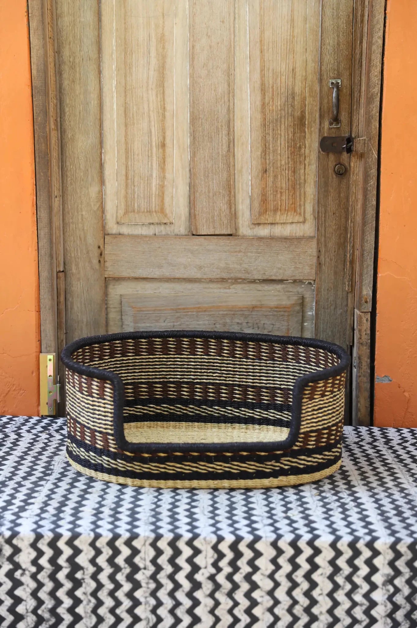 Woven pet bed with geometric pattern on a textured surface in front of a wooden door.