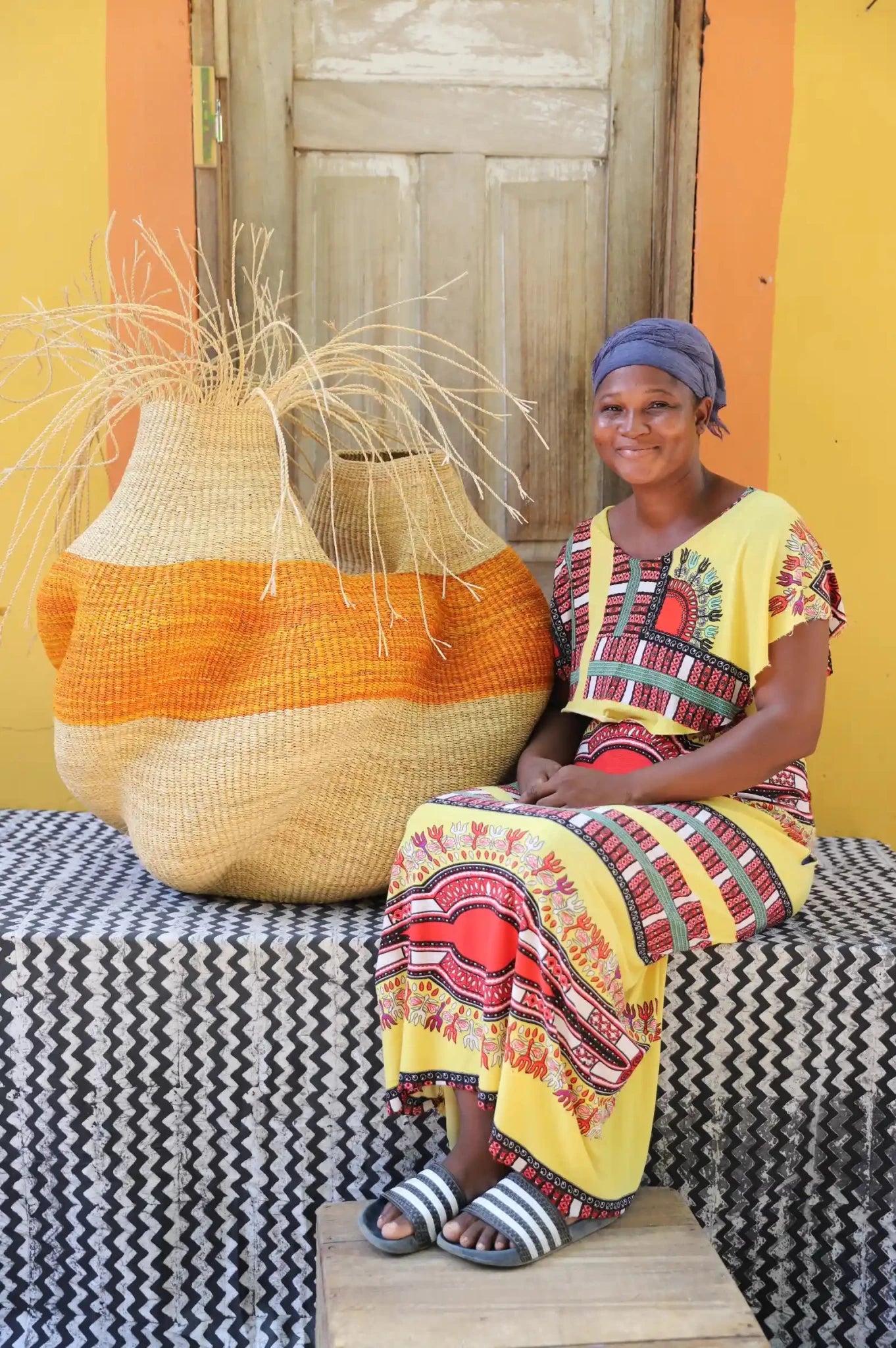 An artisan seated next to a handcrafted decorative basket with stylish straw branching out on a black and white cloth against a wooden door.