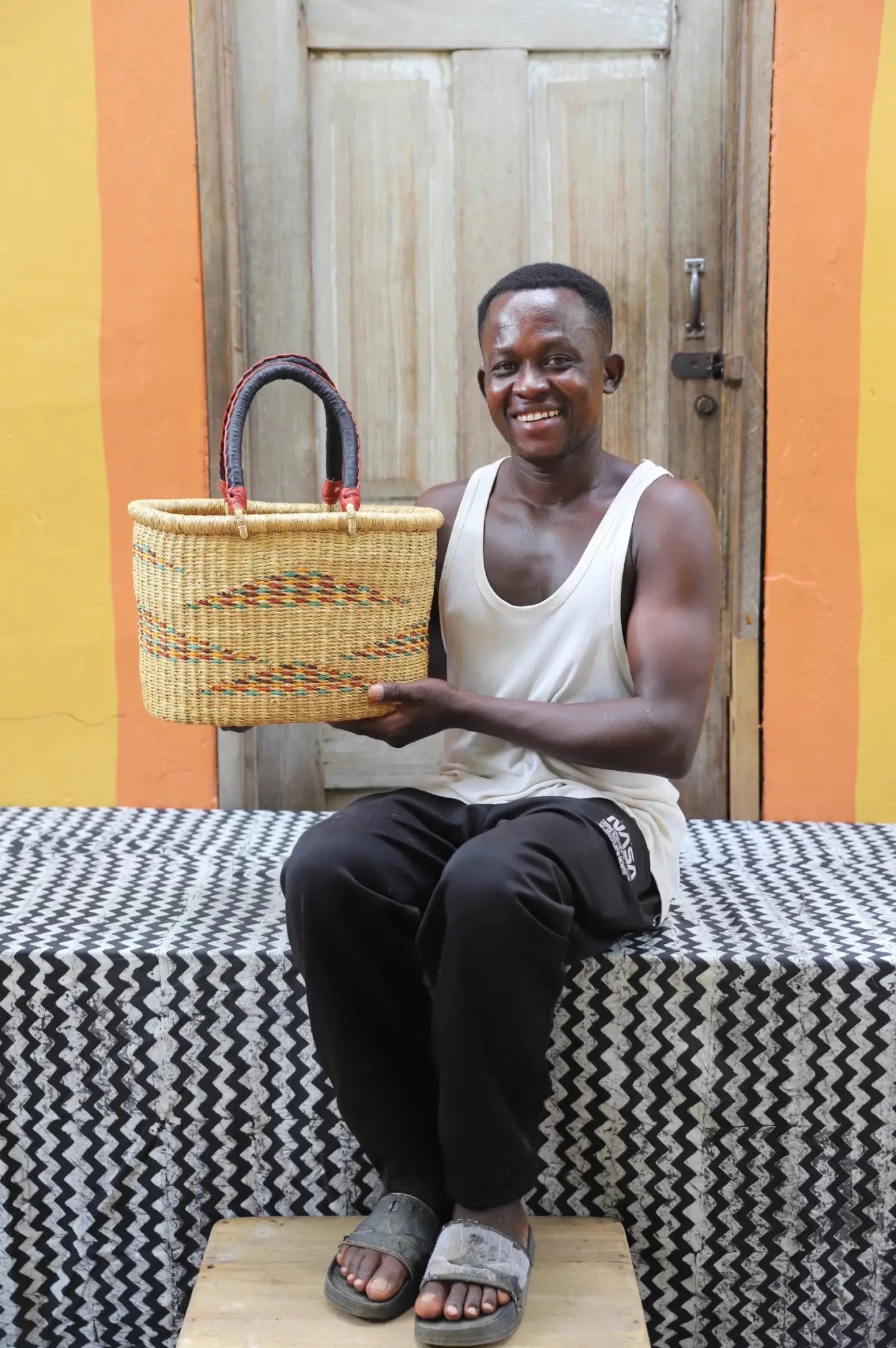 Man seated holding a medium woven carry basket in natural straw with colourful stripe detail and leather-wrapped handles