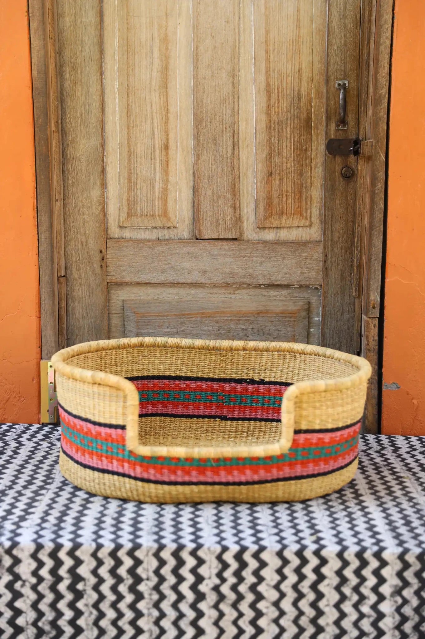 Woven pet bed with geometric pattern on a textured surface in front of a wooden door.