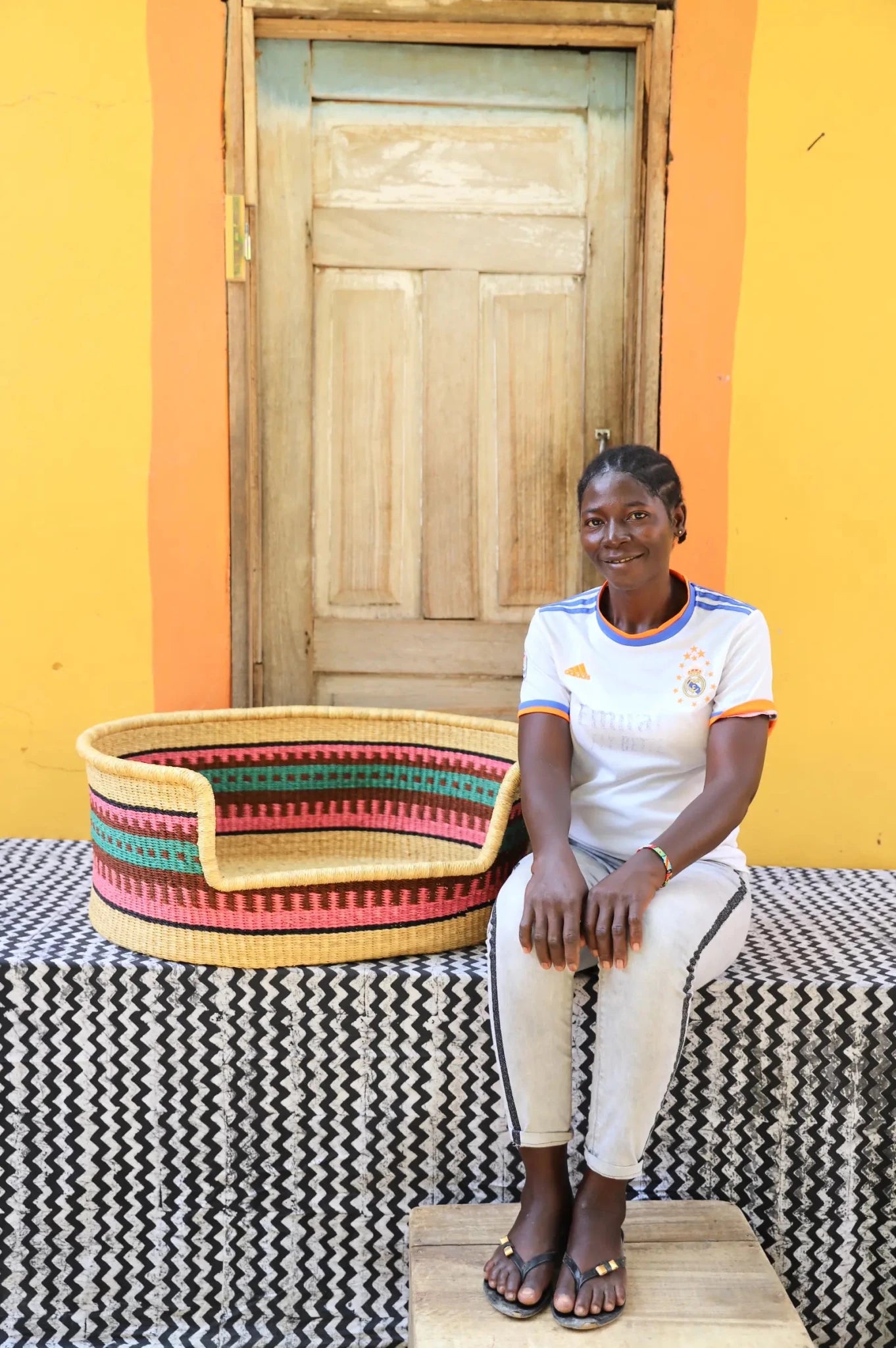 Woman  sitting on a bench with a woven basket next to a wooden door and yellow wall.