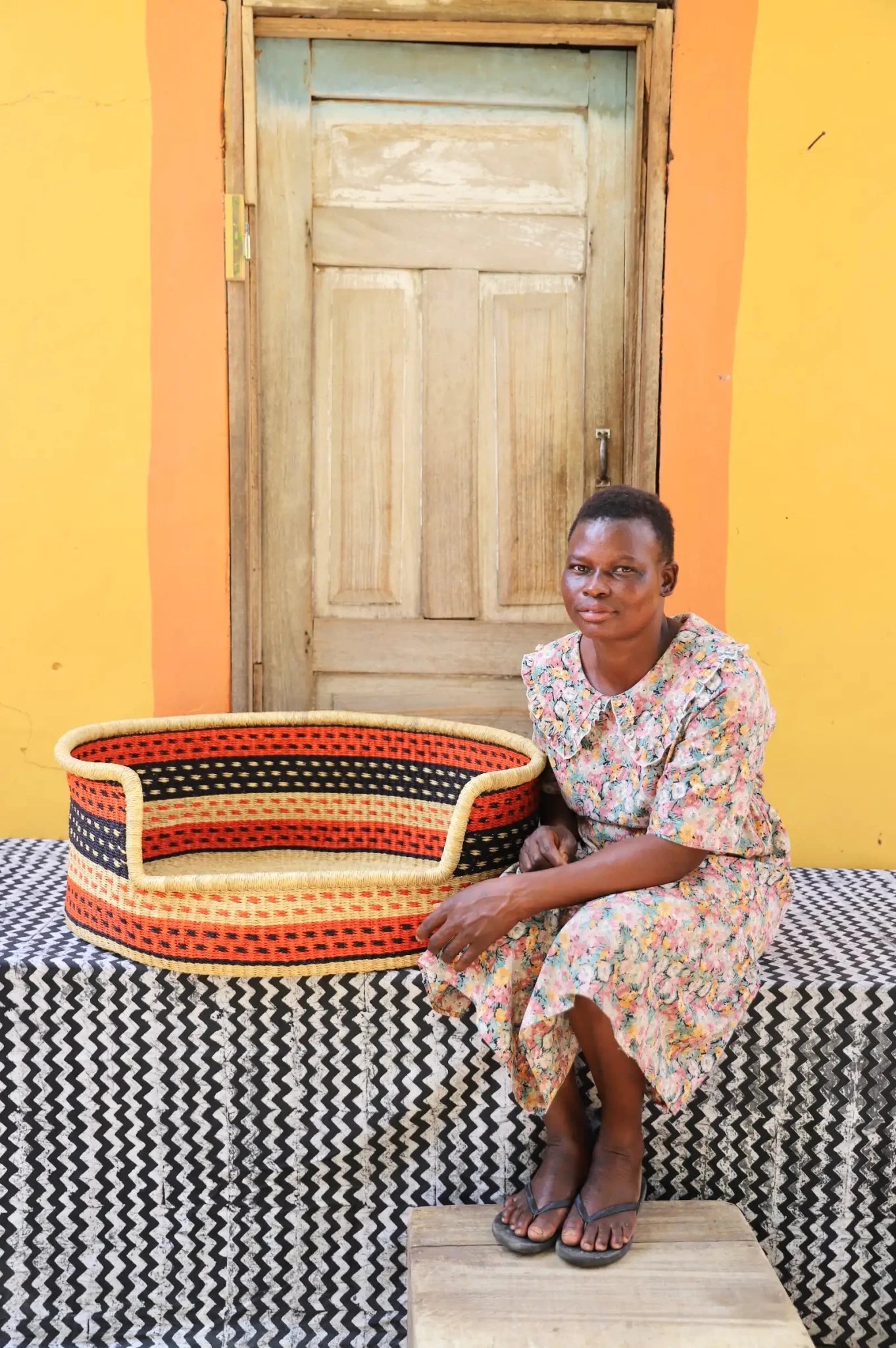 Woman sitting on a bench with a woven basket next to a wooden door and yellow wall.